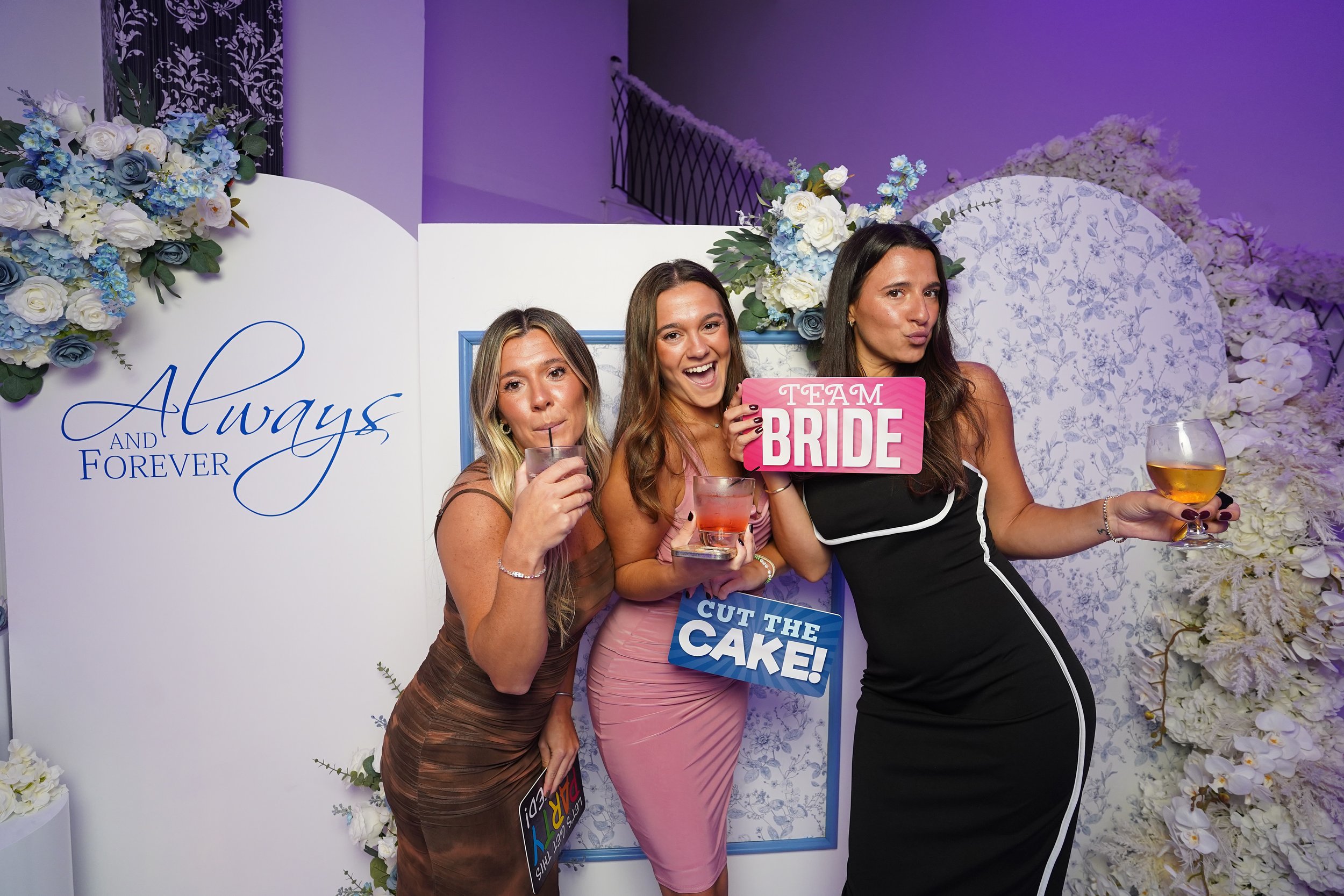 Three women at a wedding celebration posing with drinks and signs, standing in front of a floral backdrop with the words 'Always and Forever'.
