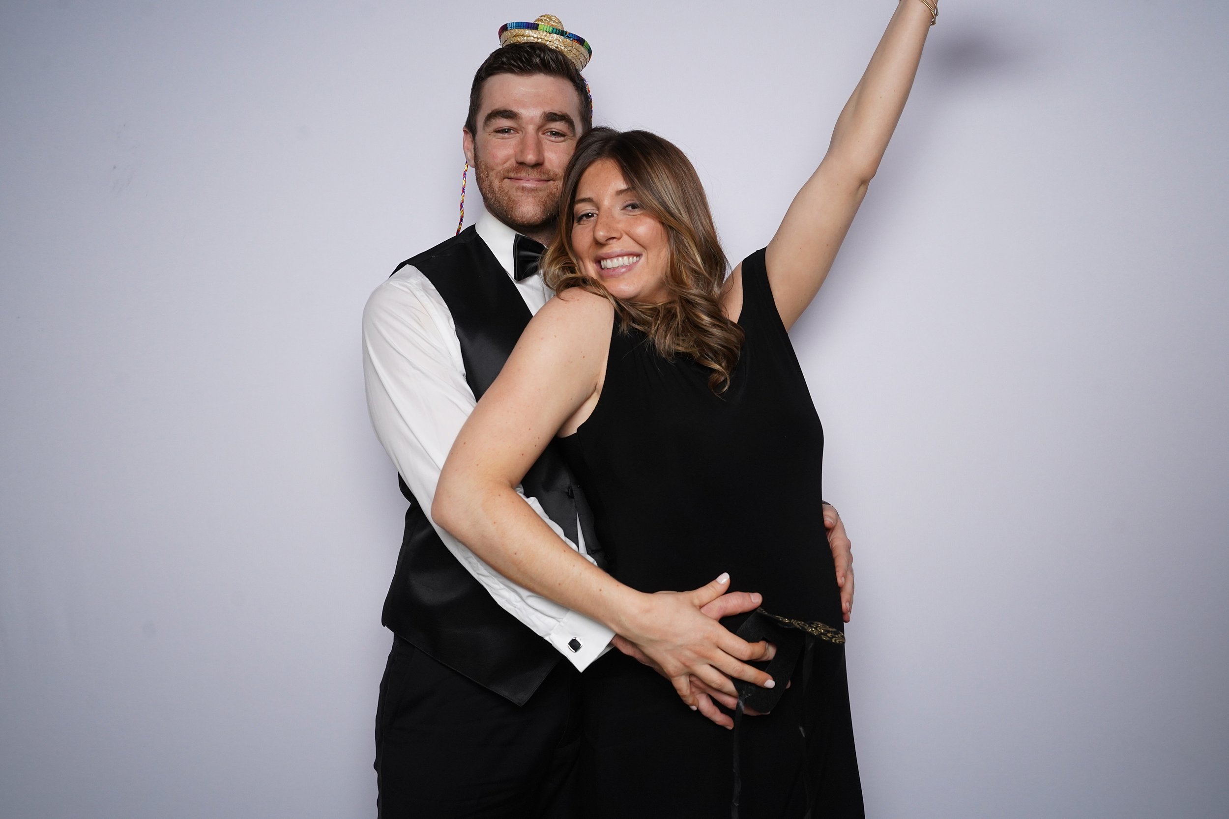 A smiling man and woman dressed formally, with the man wearing a tuxedo and the woman in a black dress, striking a playful pose against a plain background. The man has his arm around the woman, and she is raising her arm in celebration.
