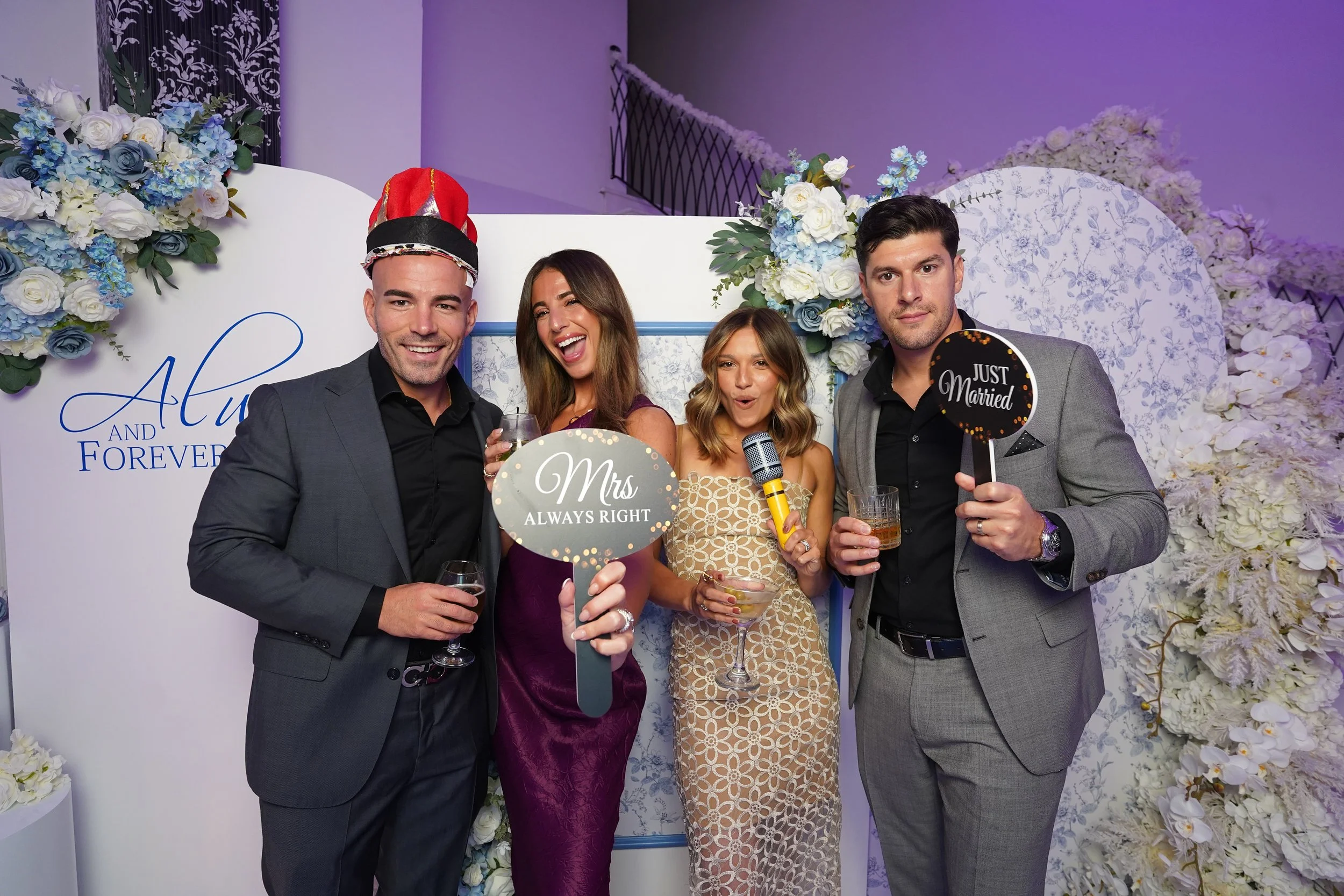 Group of four people at wedding celebration, holding signs and drinks, standing in front of decorated floral backdrop.