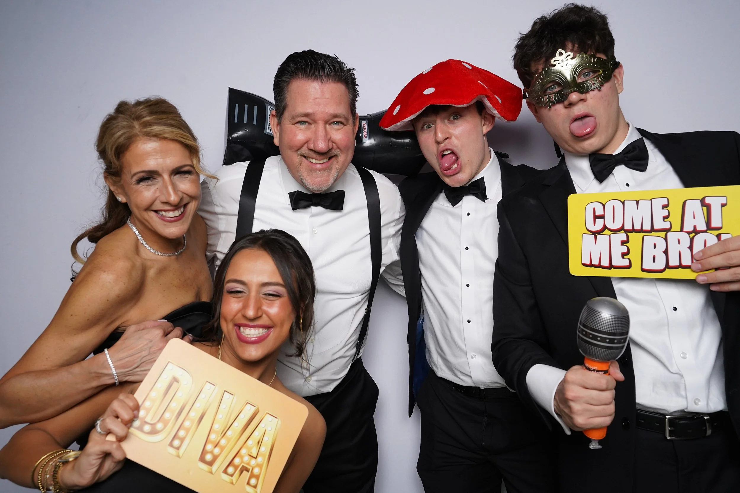 Group of five people at a party, smiling and posing for the camera, dressed in tuxedos and evening dresses, holding fun signs and props, with a plain white background.
