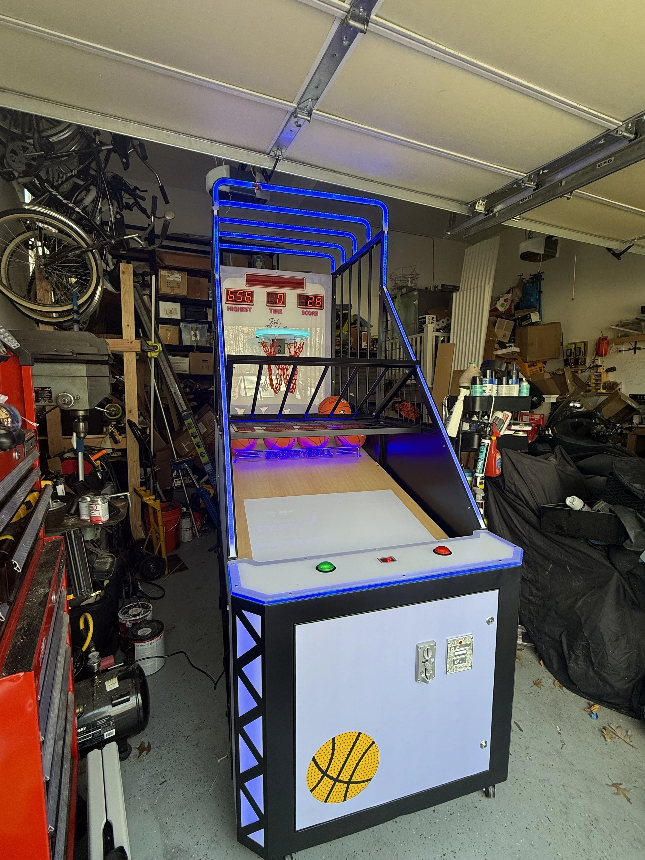 An arcade basketball game in a garage, featuring a digital score display, basketball hoops, neon blue lights, and basketballs inside the game. The garage has tools and equipment on shelves and the floor.