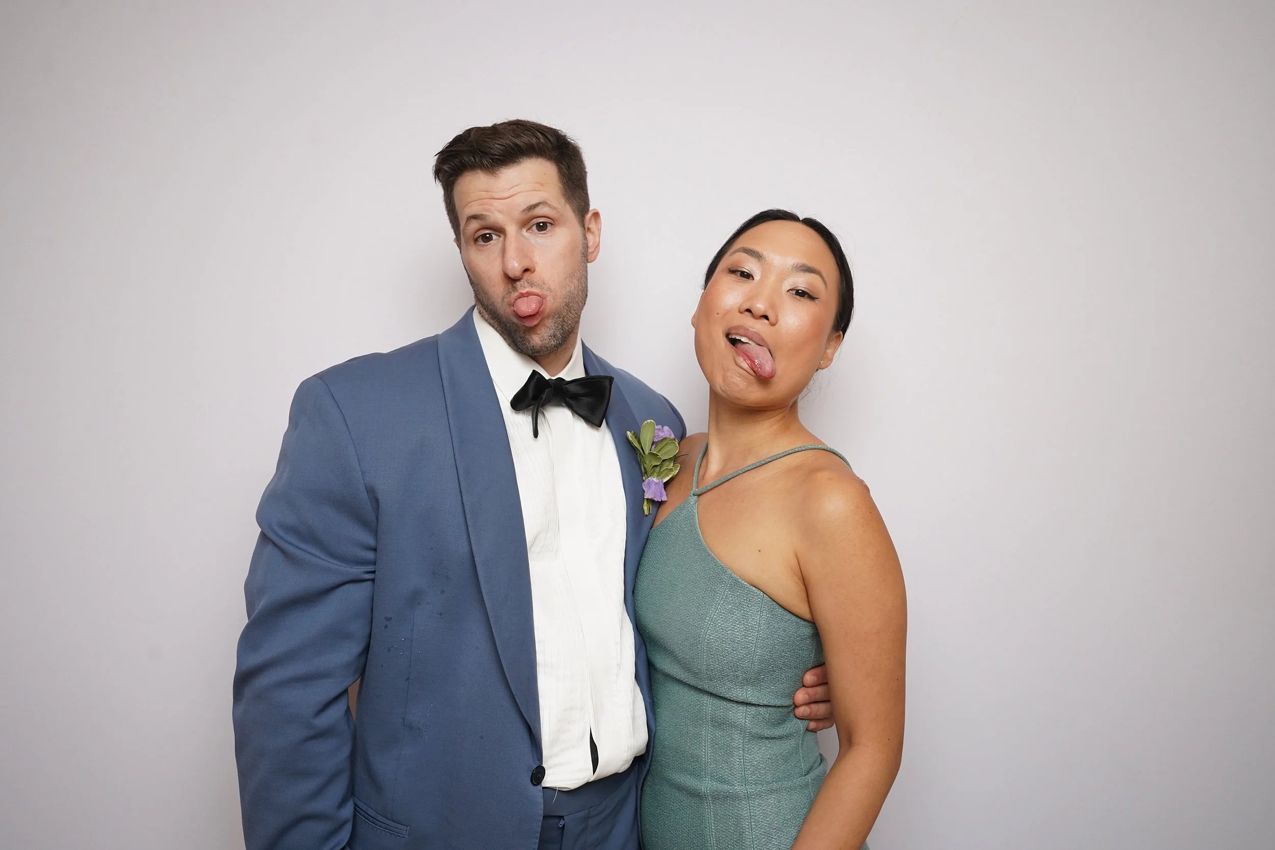 A man in a blue tuxedo and a woman in a green dress are making silly faces with their tongues out, standing close together against a plain background.