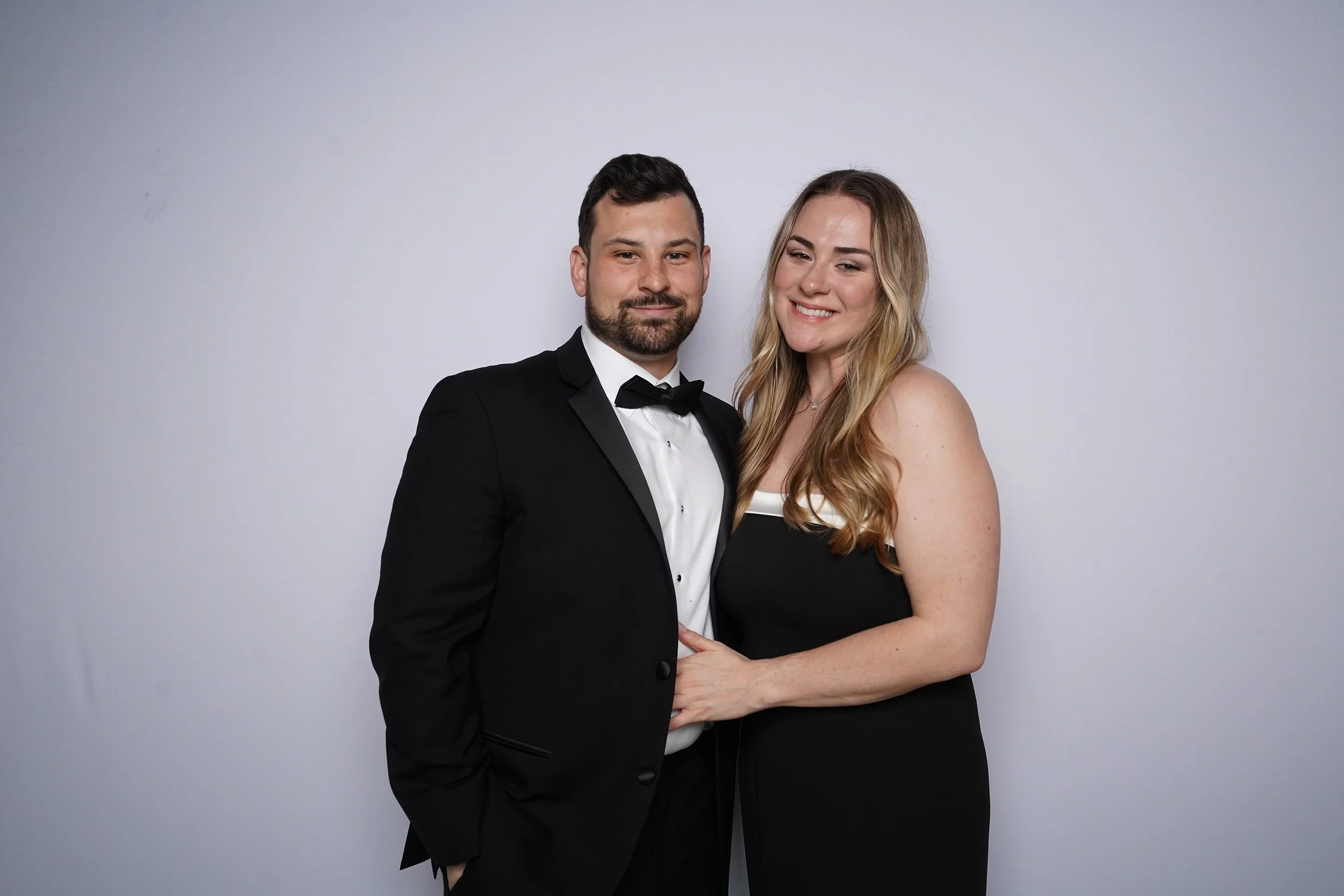 A man and woman dressed in formal attire, with the man in a black tuxedo and the woman in a black dress, standing close together smiling against a plain light gray background.