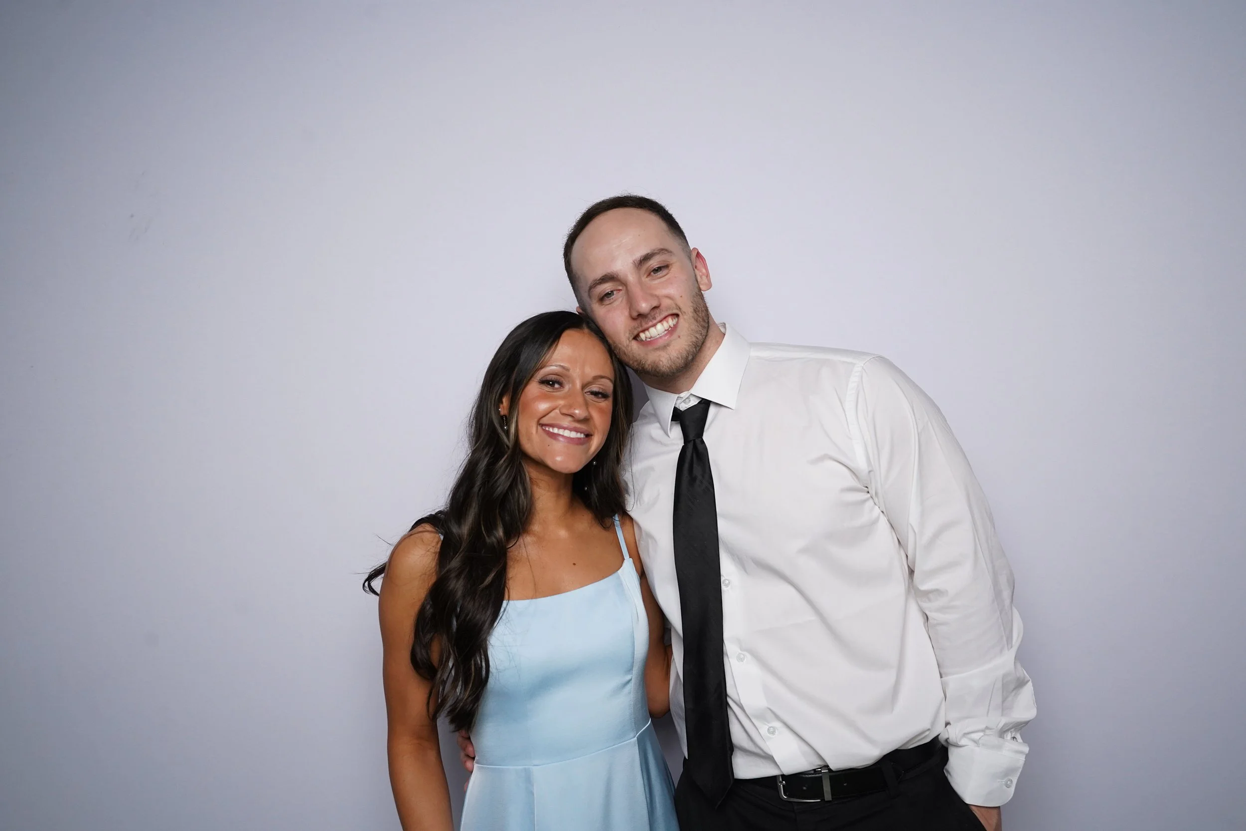 A smiling woman with long dark hair in a light blue dress stands next to a man in a white shirt and black tie. They are standing against a plain light gray background, close together with the man's arm around the woman's waist.