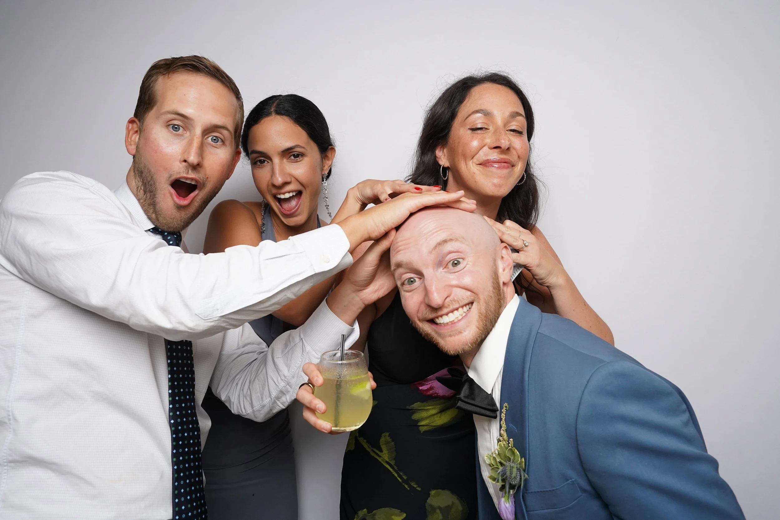 Group of four adults having fun and touching a bald man's head at a celebration, with one holding a drink, against a plain gray background.