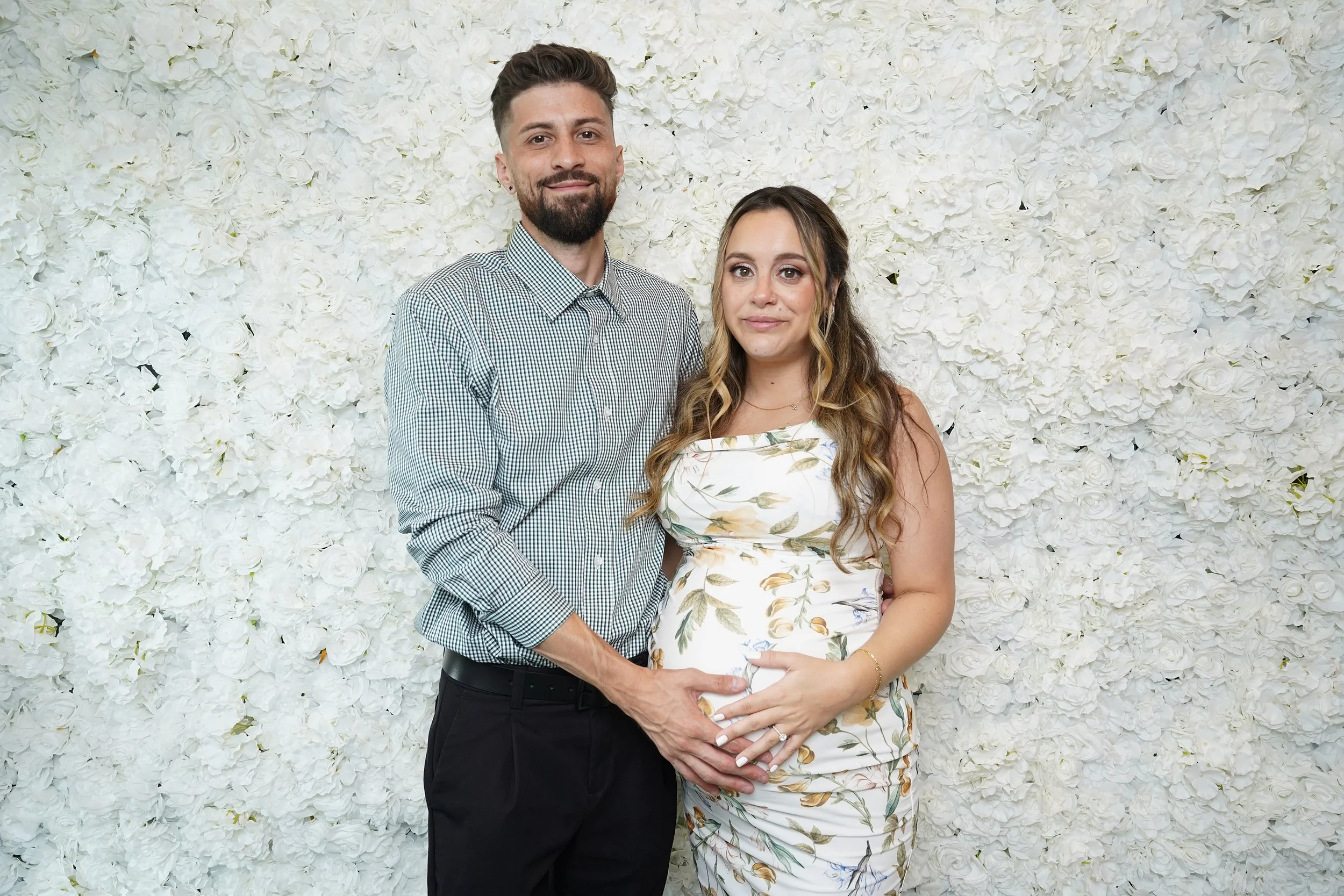 A smiling couple, with the man touching the woman's pregnant belly, standing in front of a white floral wall