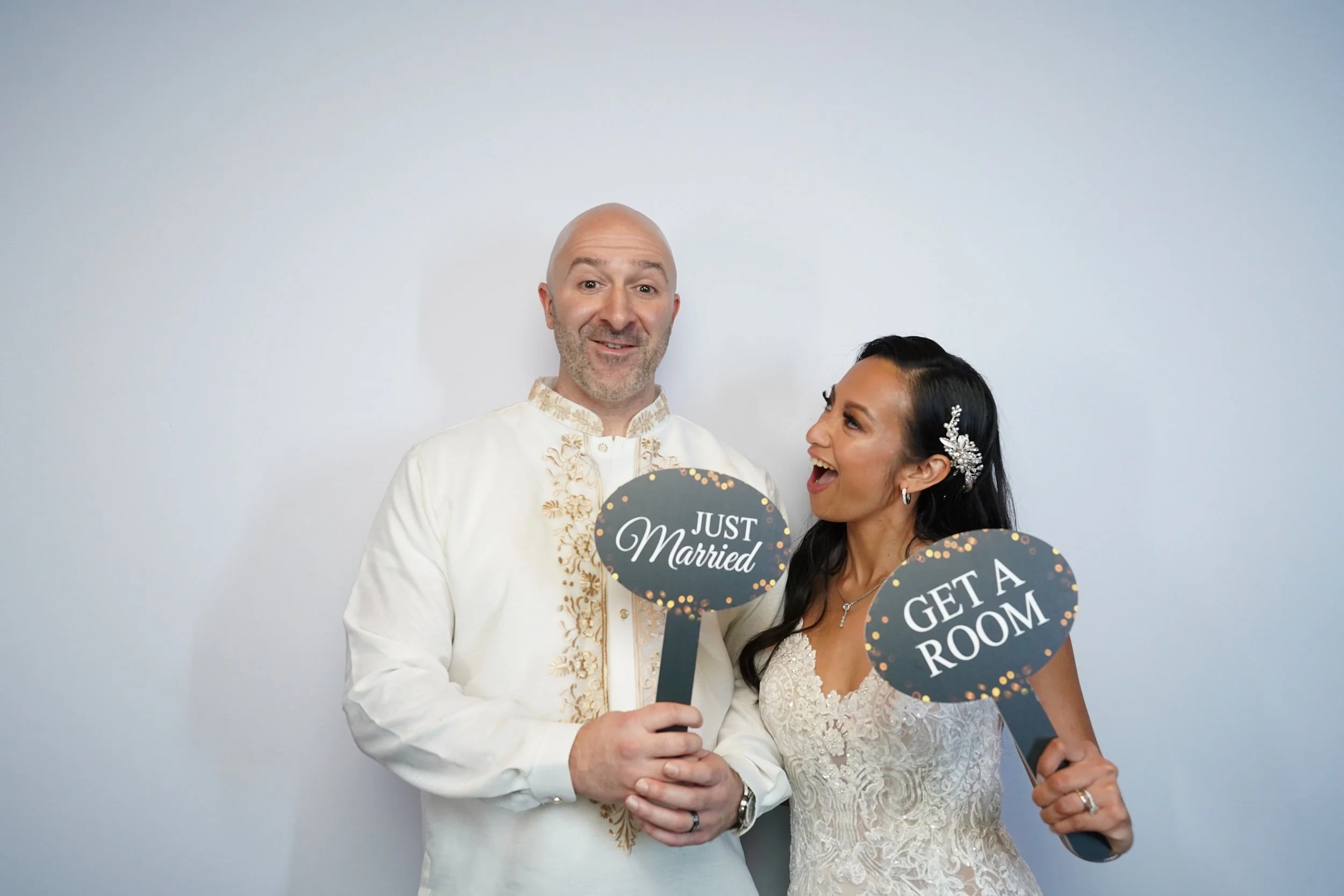 A newlywed couple dressed in wedding attire holding signs that say 'Just Married' and 'Get a Room' while smiling and having fun.