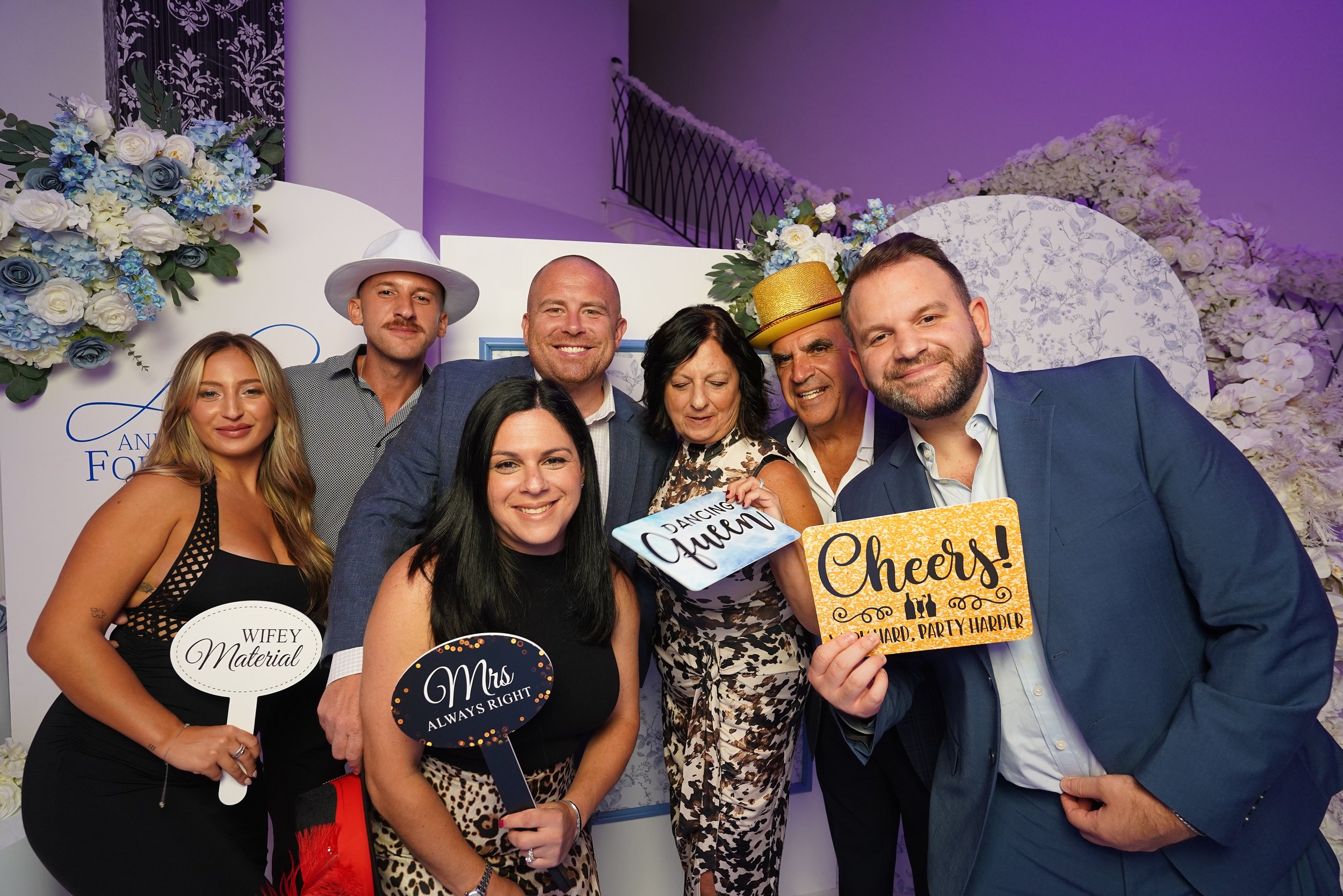 Group of eight people dressed up at a celebration, holding humorous signs and smiling, with floral decorations in the background.