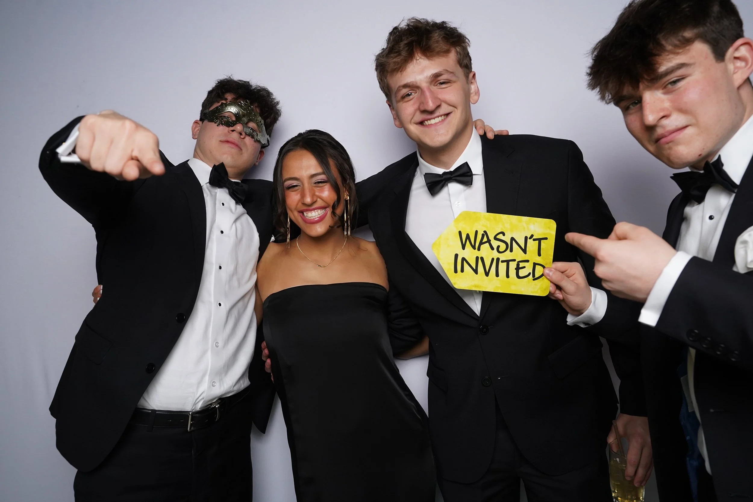 Group of four young adults at a formal event, dressed in tuxedos and a strapless black dress, posing together with smiles. One is wearing a mask, and another is holding a yellow sign that reads 'Wasn't Invited.'