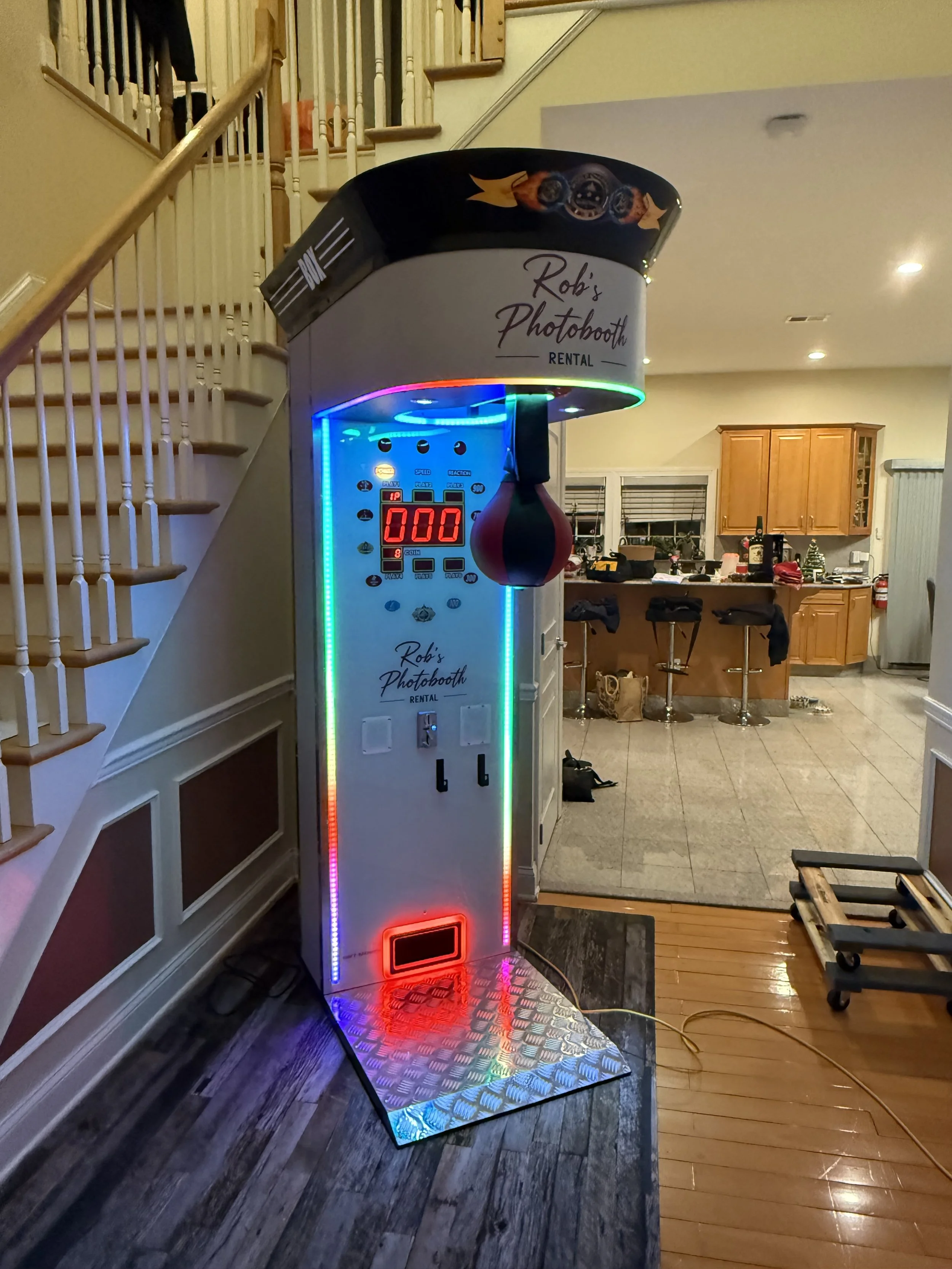 Photo booth machine labeled 'Rob's Photobooth Rental' with colorful LED lights and a punching bag hanging from the top, set up indoors in a home kitchen area.