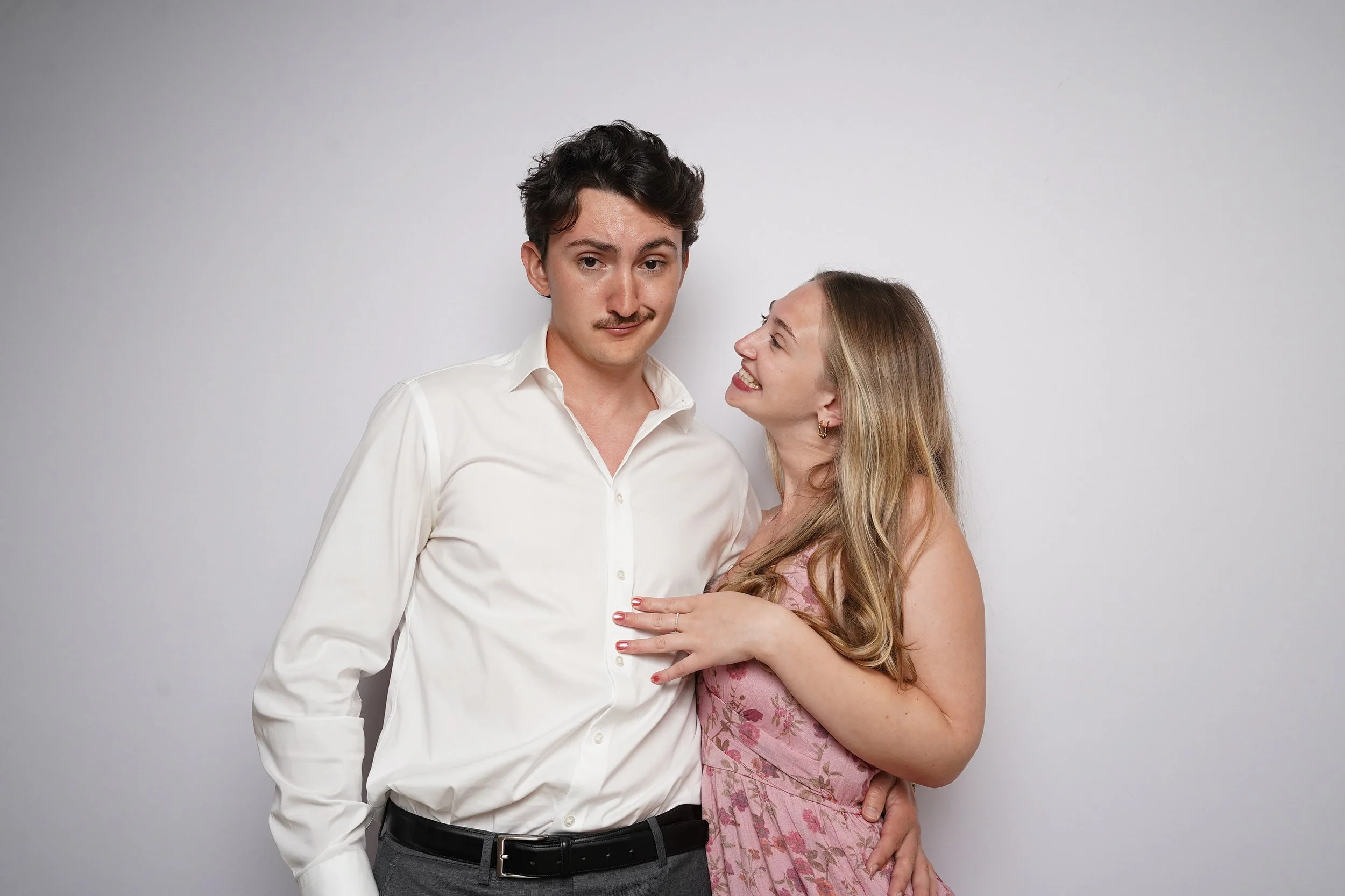 A man with dark hair and a mustache wearing a white shirt stands next to a woman with long blonde hair wearing a pink dress. The woman is smiling and looking at the man, who has a neutral expression. They are standing against a plain white wall.