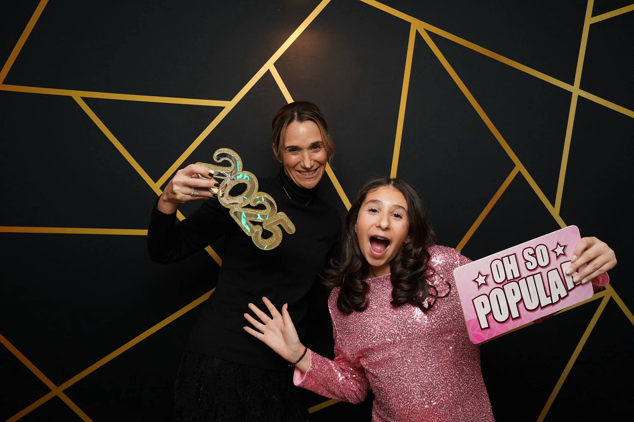 Two women celebrating New Year's Eve, one holding a sign that says 'OH SO POPULAR' and the other holding a glittery '2025' decoration, in front of a black and gold geometric background.