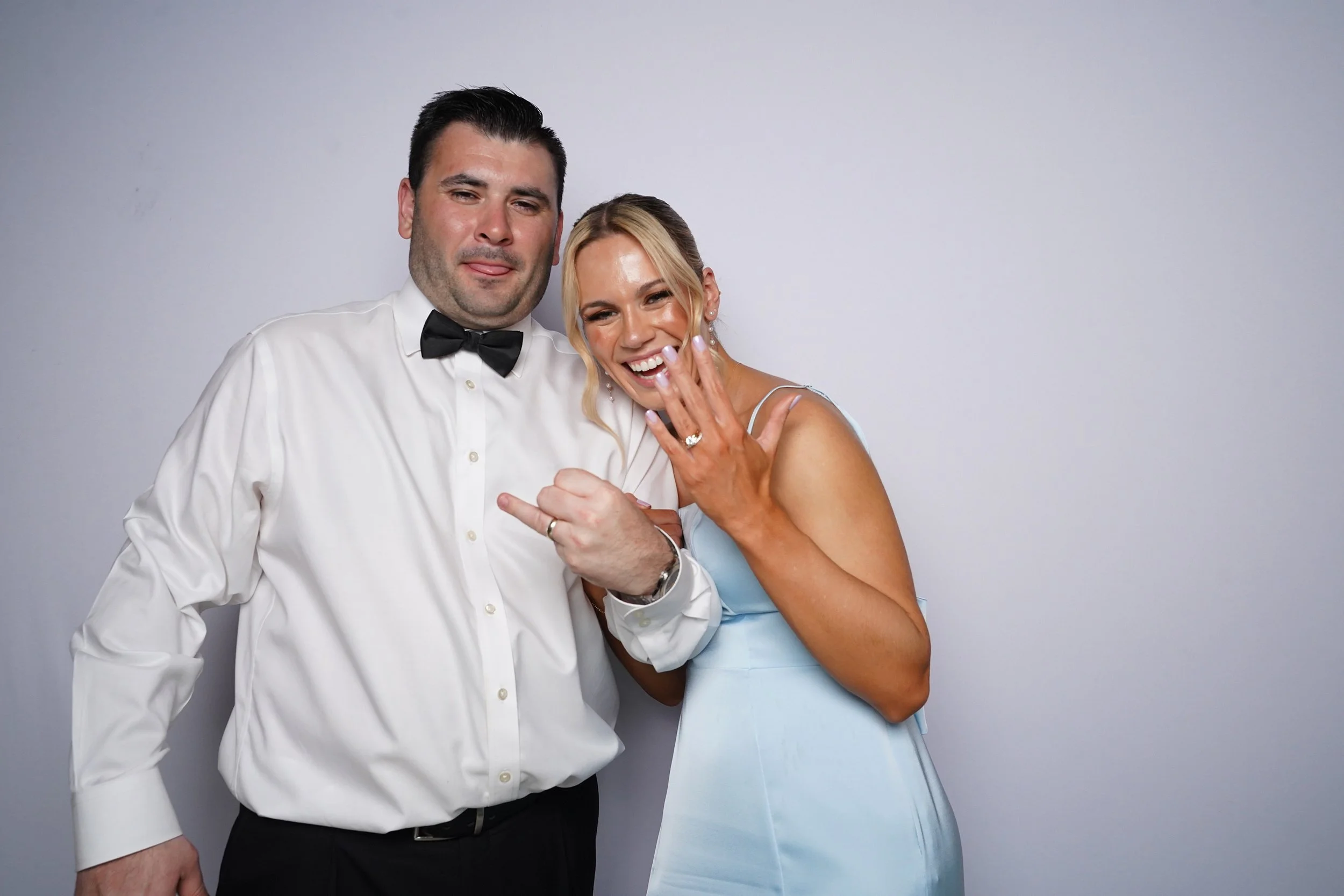 A couple dressed formally, the man in a white shirt with a black bow tie and the woman in a light blue dress, posing together and smiling at a photo booth.