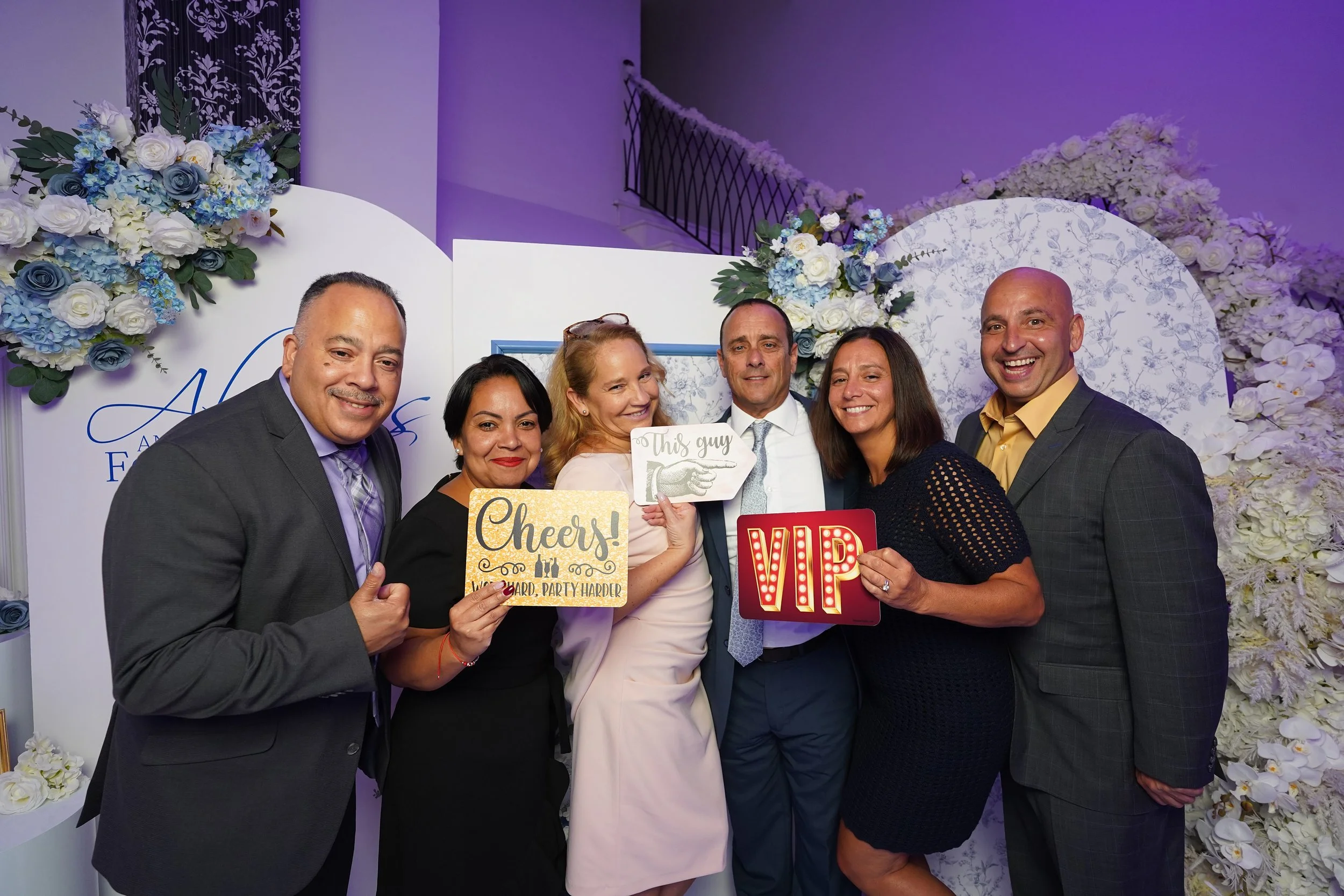 Group of six people at a celebration, holding signs that say chances, VIP, and this guy, with smiles and flowers in background.