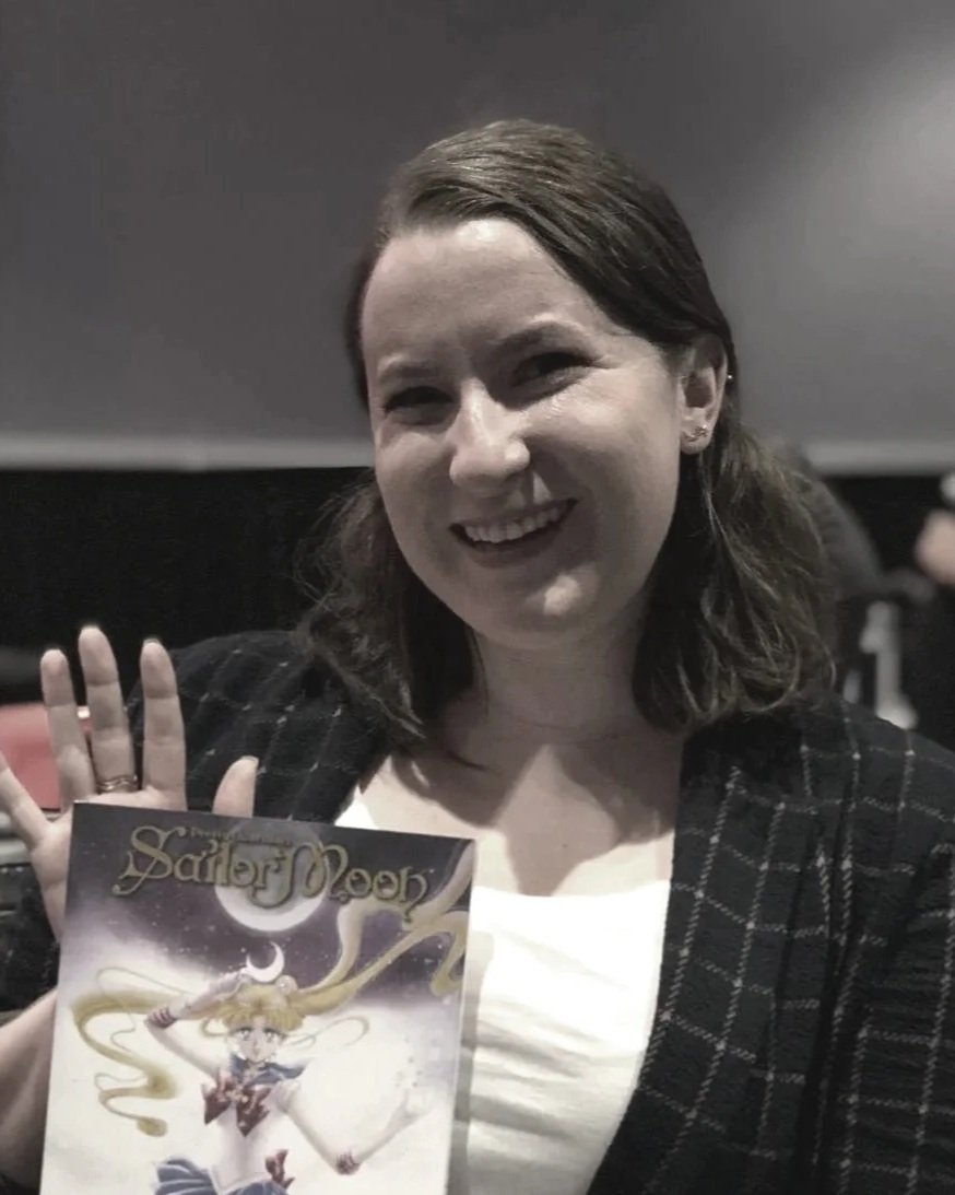 A woman smiling and waving, holding a comic book titled "Sailor Moon" in her left hand, indoors with a dark background.