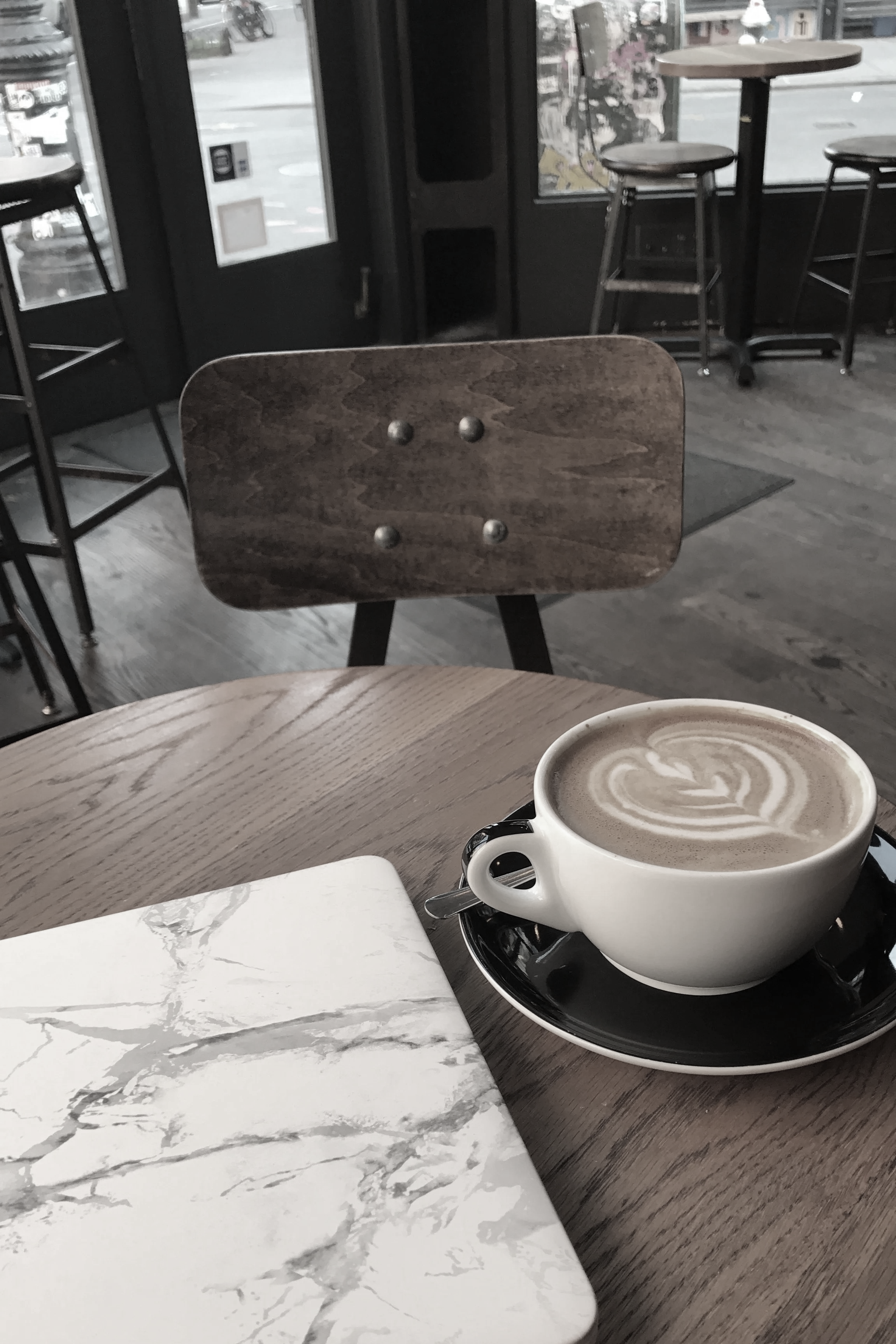 A cup of coffee with latte art on a wooden table in a cafe, with a newspaper or magazine nearby.