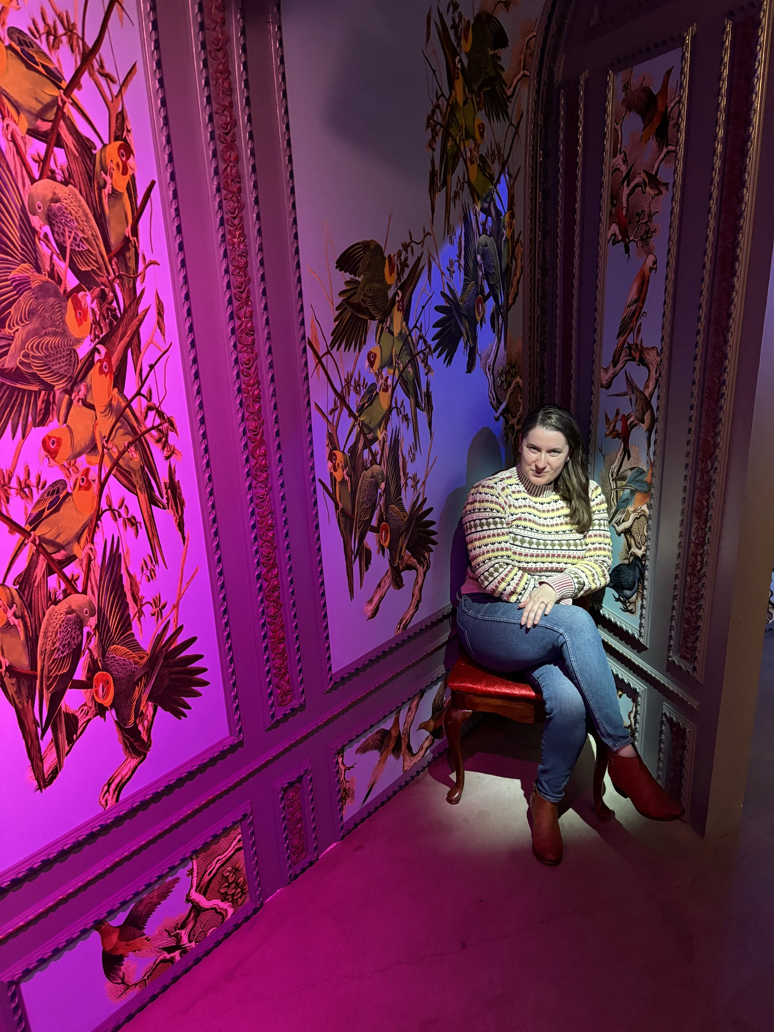 A woman sitting on a red chair in front of ornately decorated pink and purple wall panels with bird and floral illustrations, illuminated by pink and purple lighting.