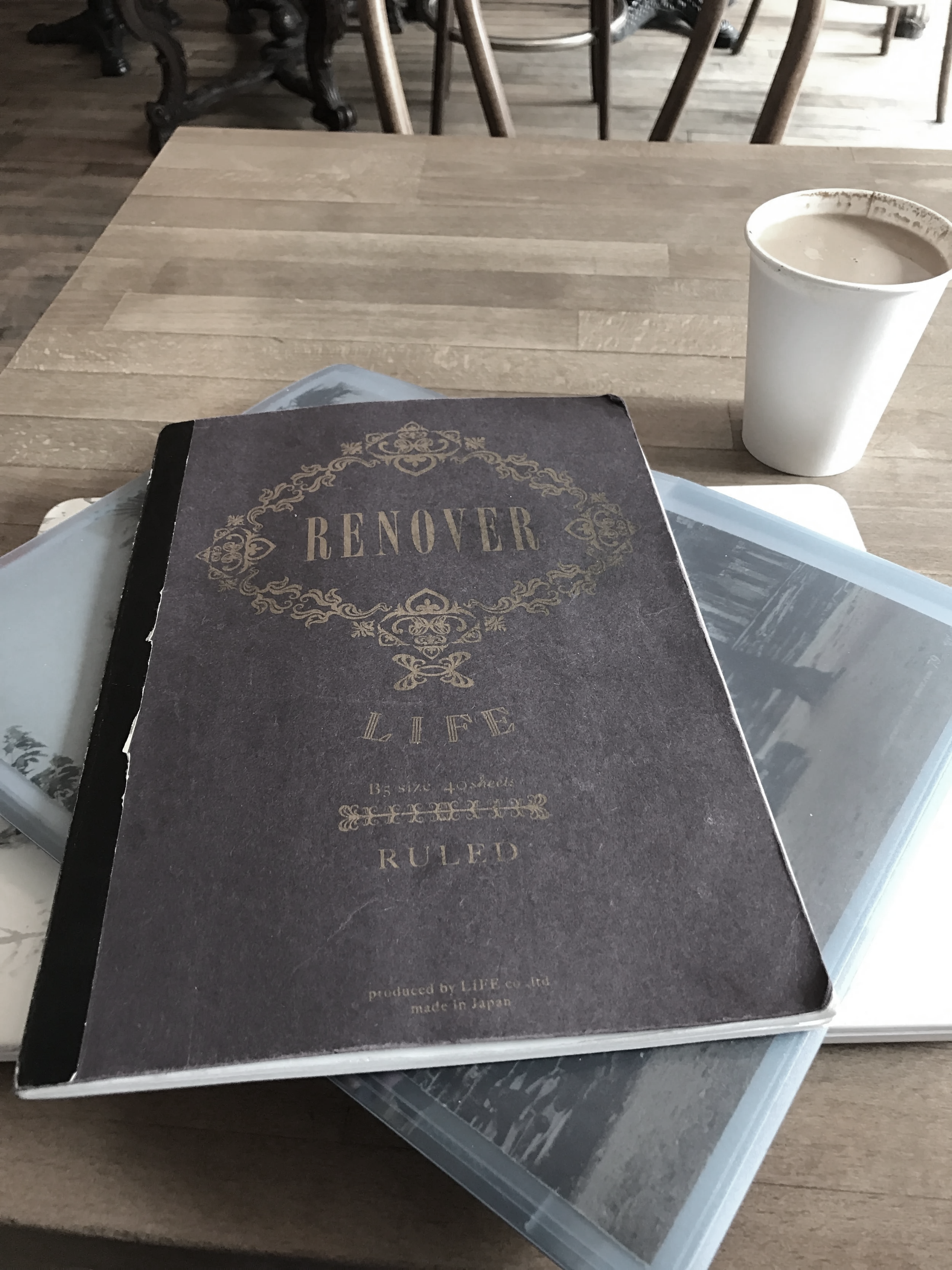 A table with a book titled 'REOVER LIFE RULED' and a white cup filled with a beverage. There are also some magazines or photo books underneath the book, in a cozy setting.
