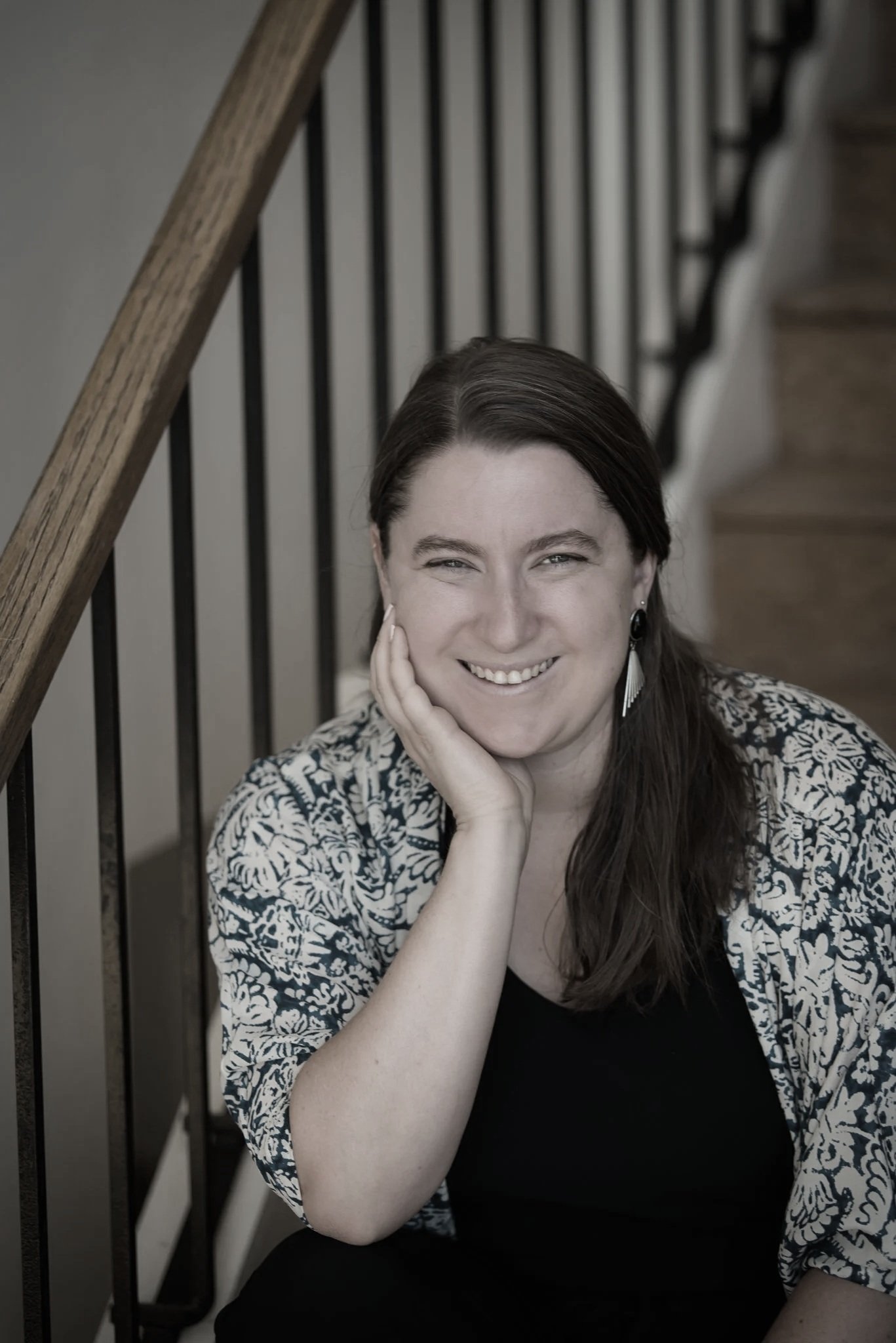 A woman with brown hair, smiling and resting her face on her hand, sitting on stairs next to a wooden and metal railing.