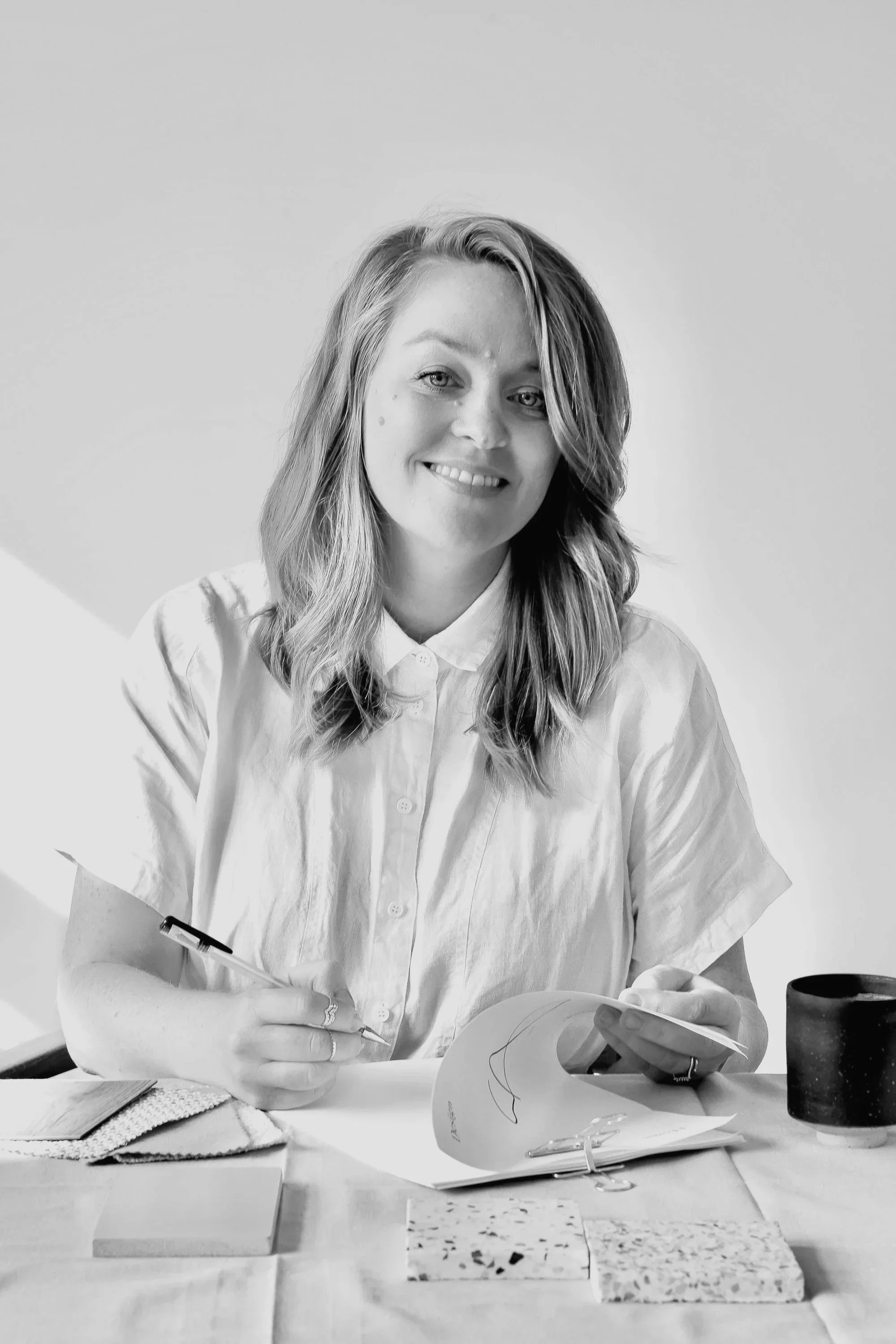 A woman with shoulder-length hair, wearing a light-colored button-up shirt, sitting at a table with various objects, holding a paper and a pen, smiling at the camera.