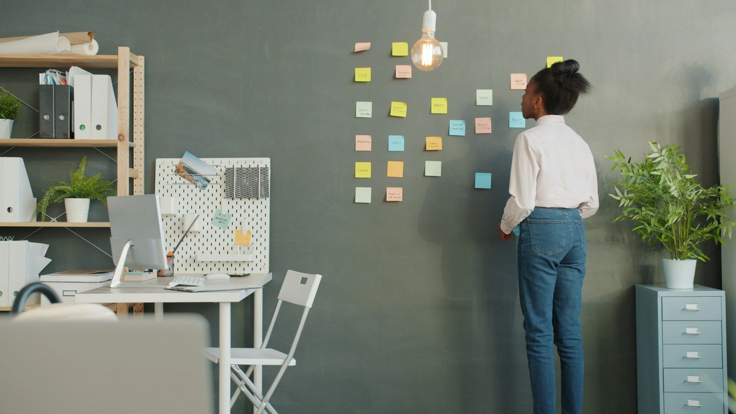 Woman standing in an office, looking at a wall with colorful sticky notes arranged in columns. There is a desk with a computer and office supplies on the left, and a shelving unit with binders and plants on the left. A lightbulb hangs from the ceiling.