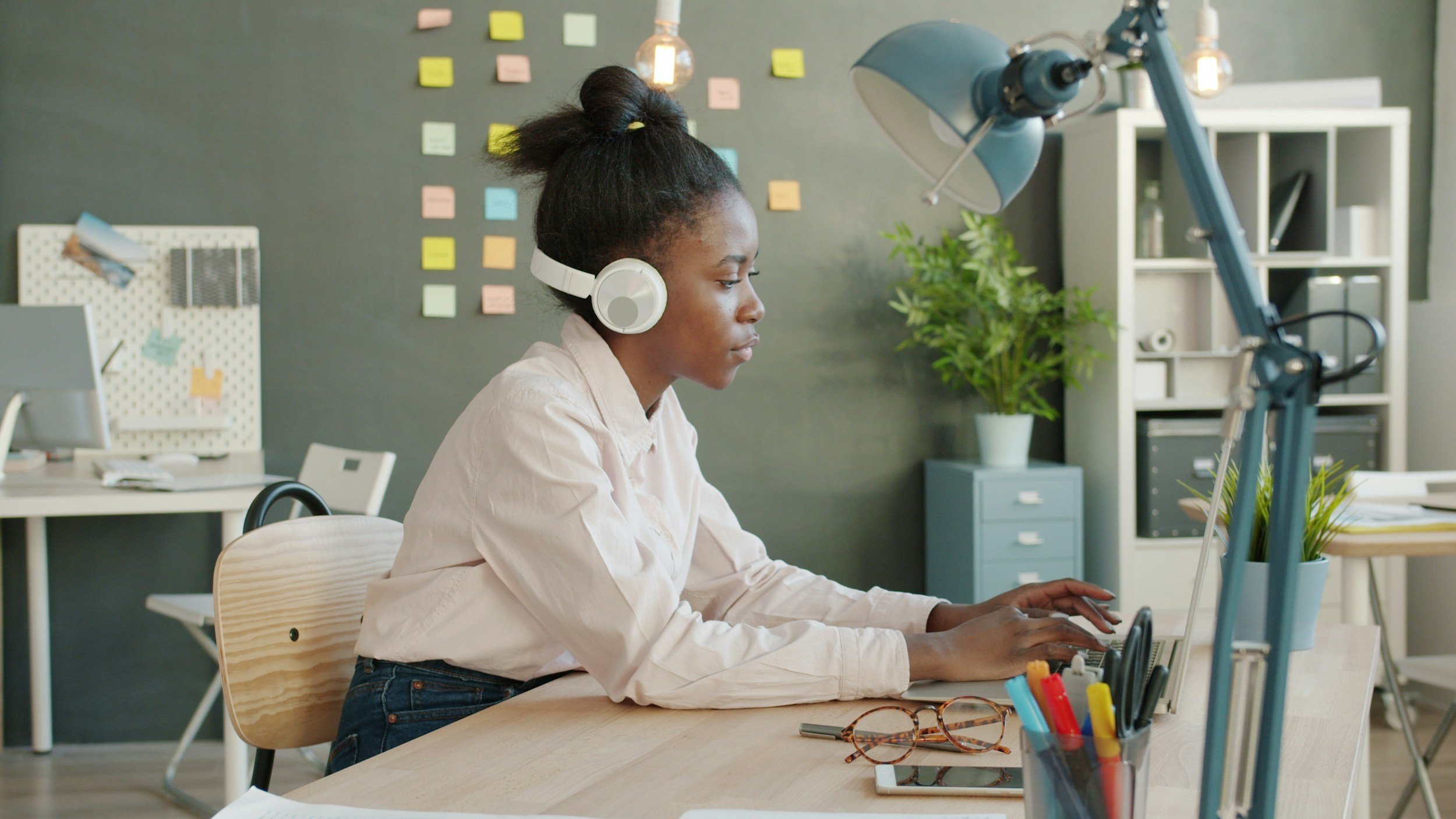 A woman sitting at a desk working on a laptop with a pen holder, glasses, and a smartphone nearby in a modern office