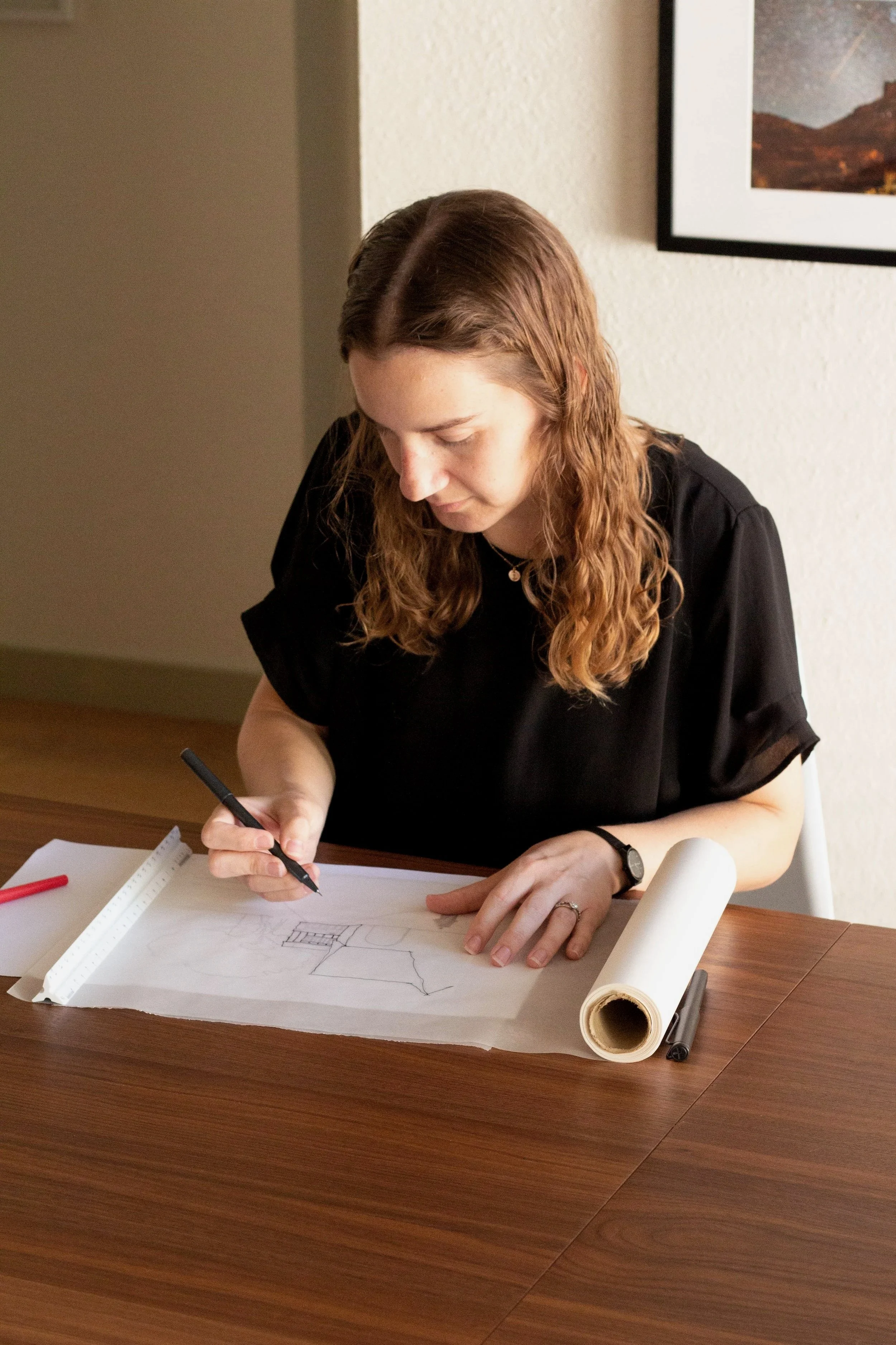 A woman with wavy, light brown hair sitting at a wooden table, drawing architectural plans on blueprints with a black marker.