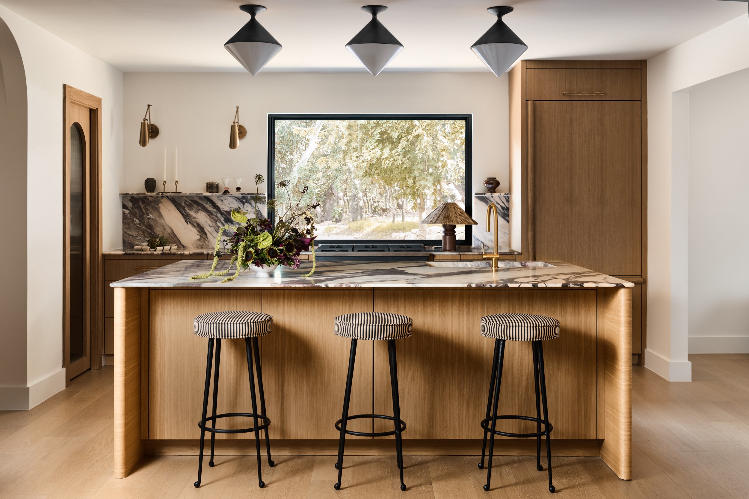 Modern kitchen with a marble island, three bar stools, a window overlooking a green outdoor area, and wooden cabinetry.