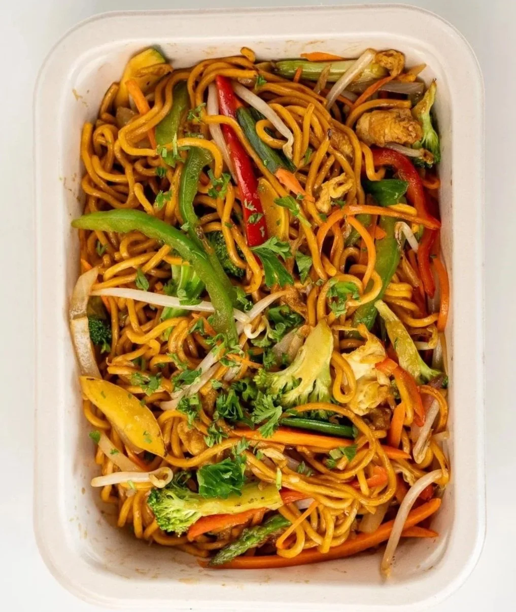 A rectangular white container filled with stir-fried noodles and mixed vegetables, including broccoli, bell peppers, carrots, and bean sprouts.
