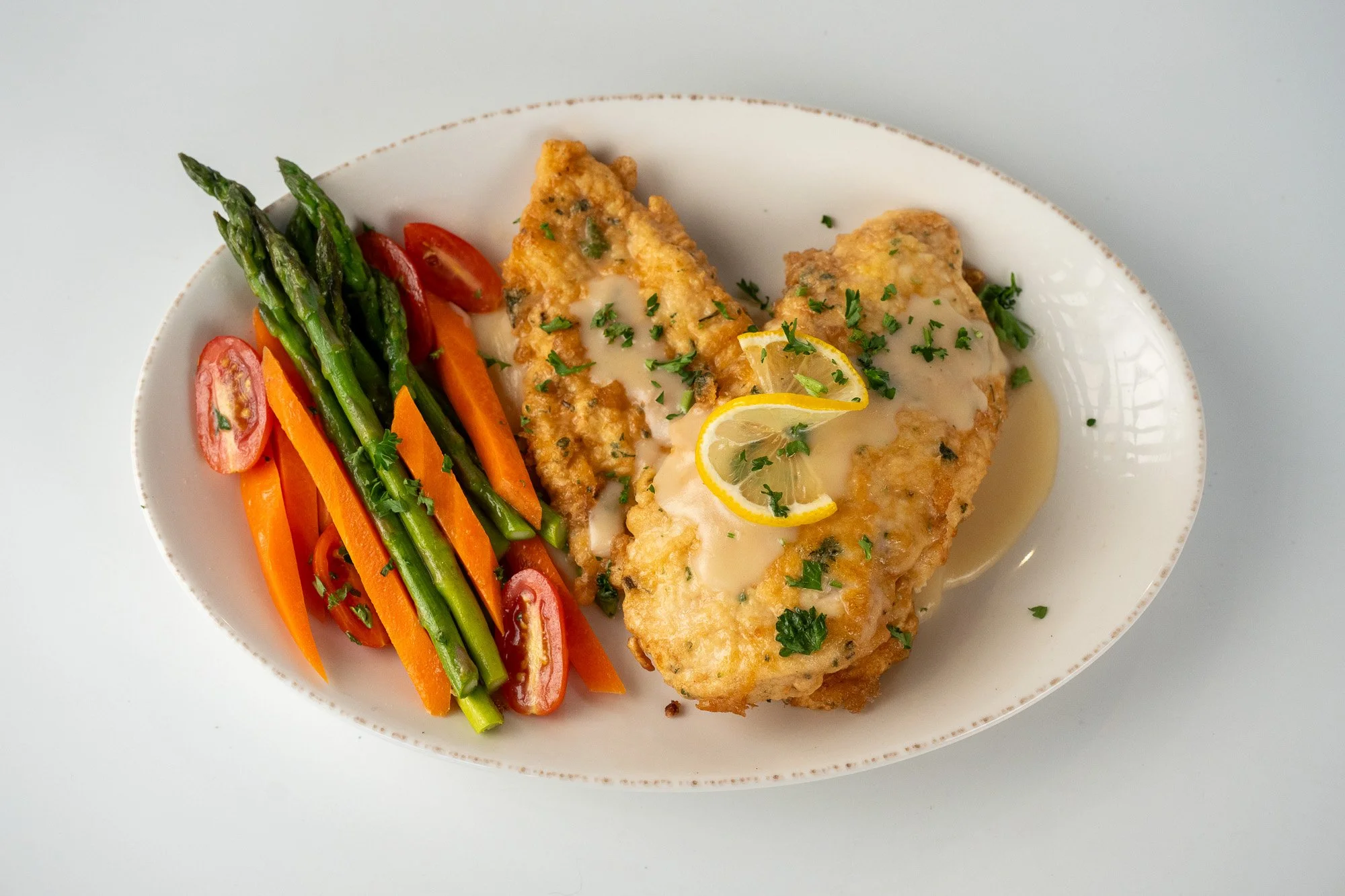 Breaded fish fillets with lemon and parsley sauce, served with sautéed carrots, asparagus, and cherry tomatoes.