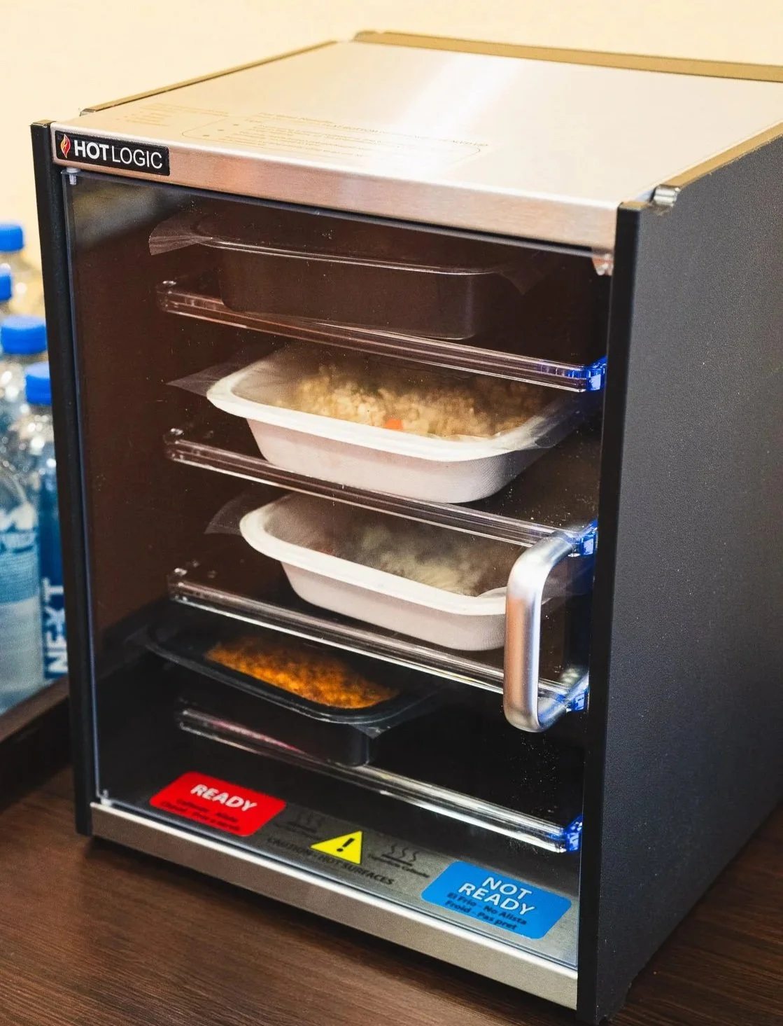 Small oven with transparent front door containing two bowls of food being heated, and warning labels on the bottom indicating ready and not ready.