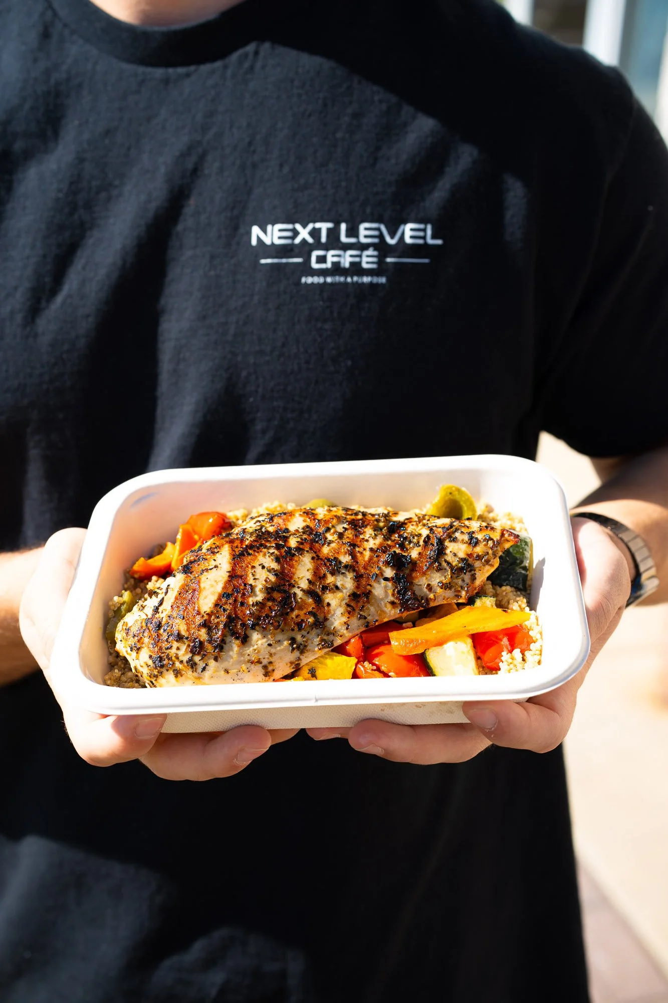 A person wearing a black shirt with 'Next Level Café' written on it, holding a white container with grilled fish and mixed vegetables.