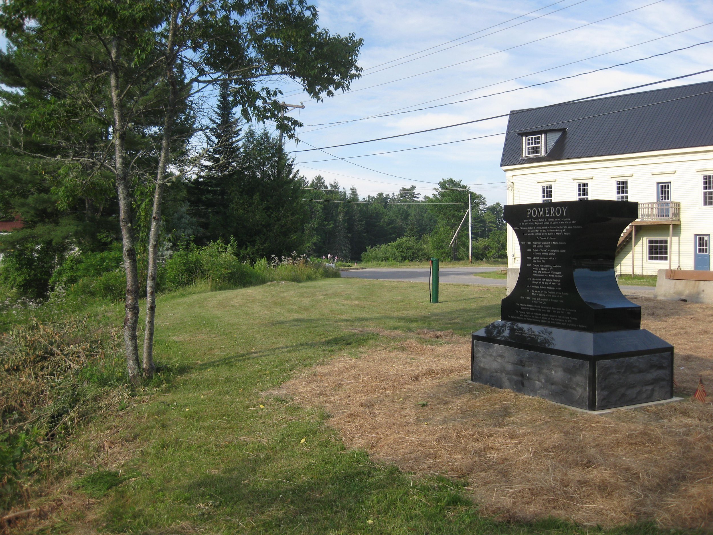 A grassy area with trees on the left, and a white building with a black roof on the right. There is a shiny black memorial or monument with the word "POMEROY" on it, situated near the building.