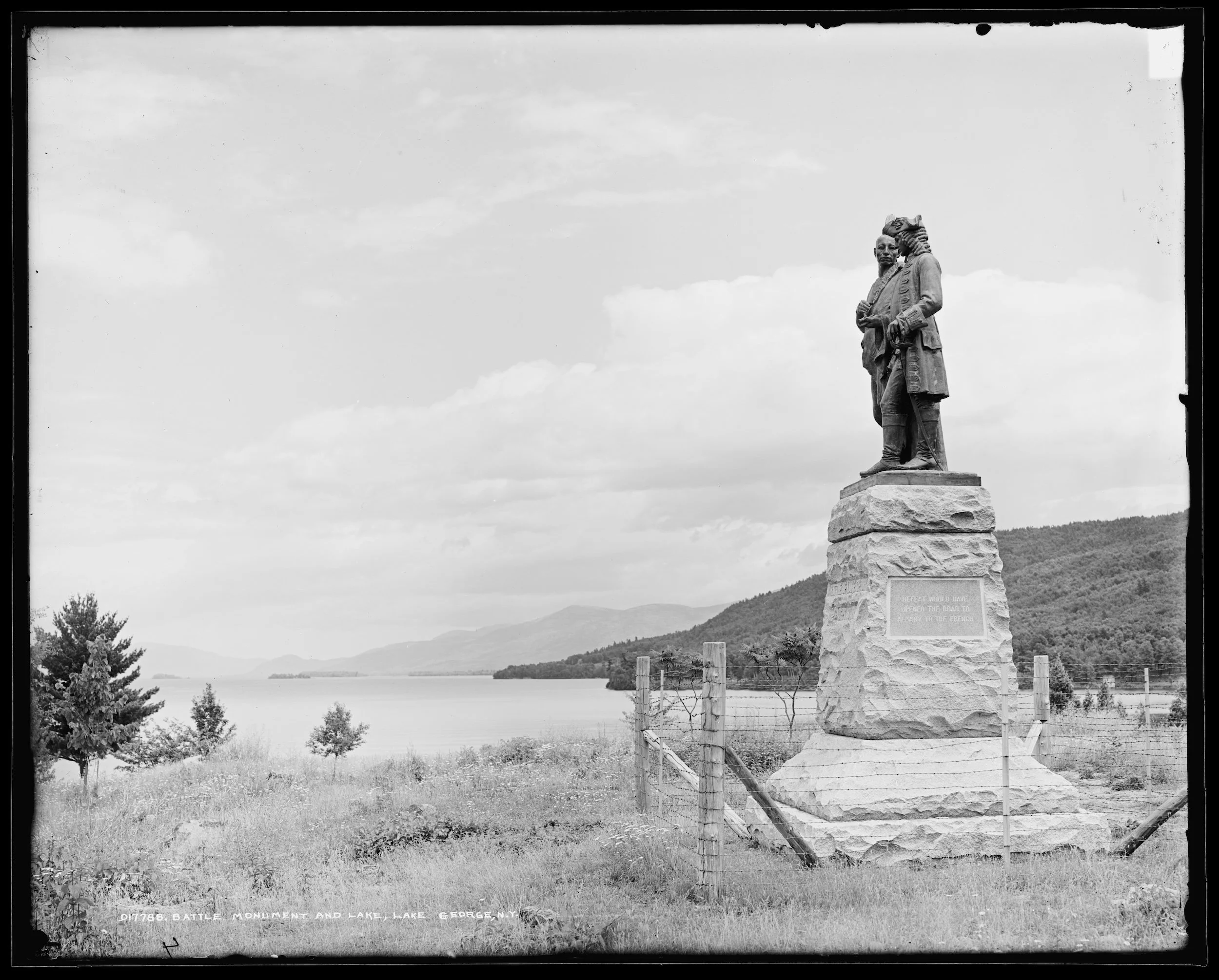 A black and white photo of a stone monument with a statue of two men, located near a lake with mountains in the background. The monument has an inscription on the front.
