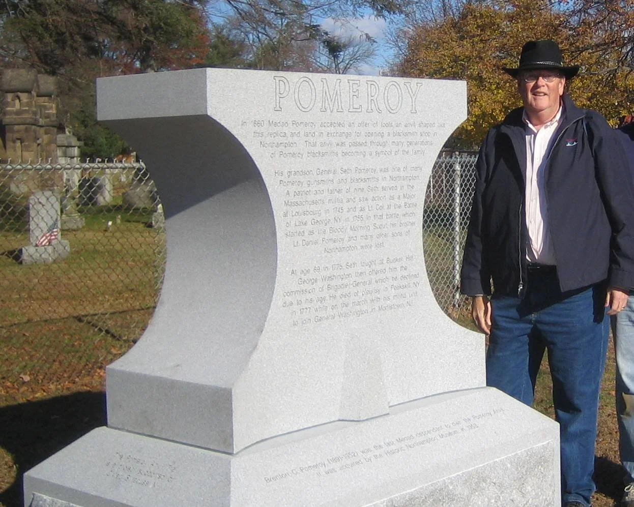 Man wearing sunglasses, a black hat, a black jacket, and blue jeans standing next to a gray monument with inscription 'Pomeroy', in a cemetery with trees and gravestones in the background.