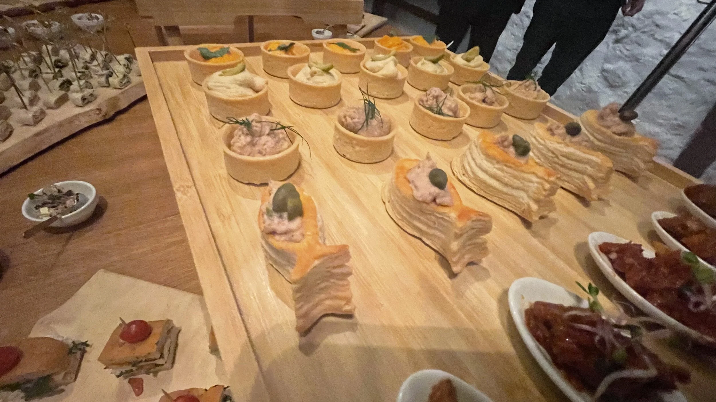 Verschiedene kleine Häppchen und Vorspeisen auf einem hölzernen Serviertablett, umgeben von weiteren kleinen Schälchen mit Speisen.