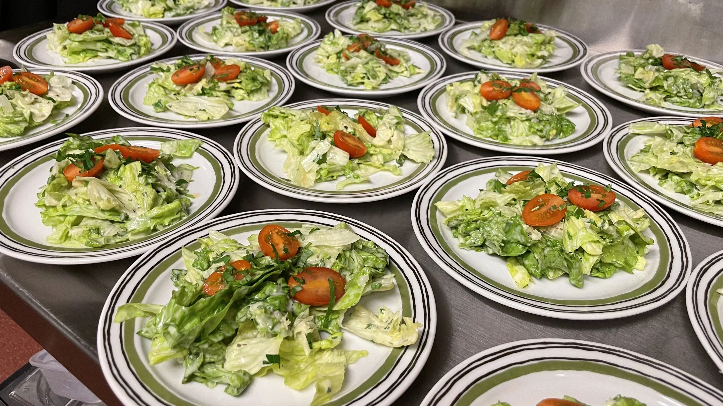 Mehrere Teller mit Salat aus Salatblättern, Cherrytomaten und gehackten Kräutern, auf einem dunklen Tisch.