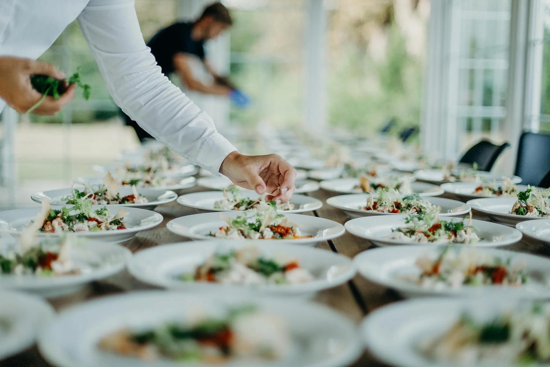 Catering-Personen bereiten mehrere Teller mit Salat in einem hellen Raum mit grossen Fenstern vor.