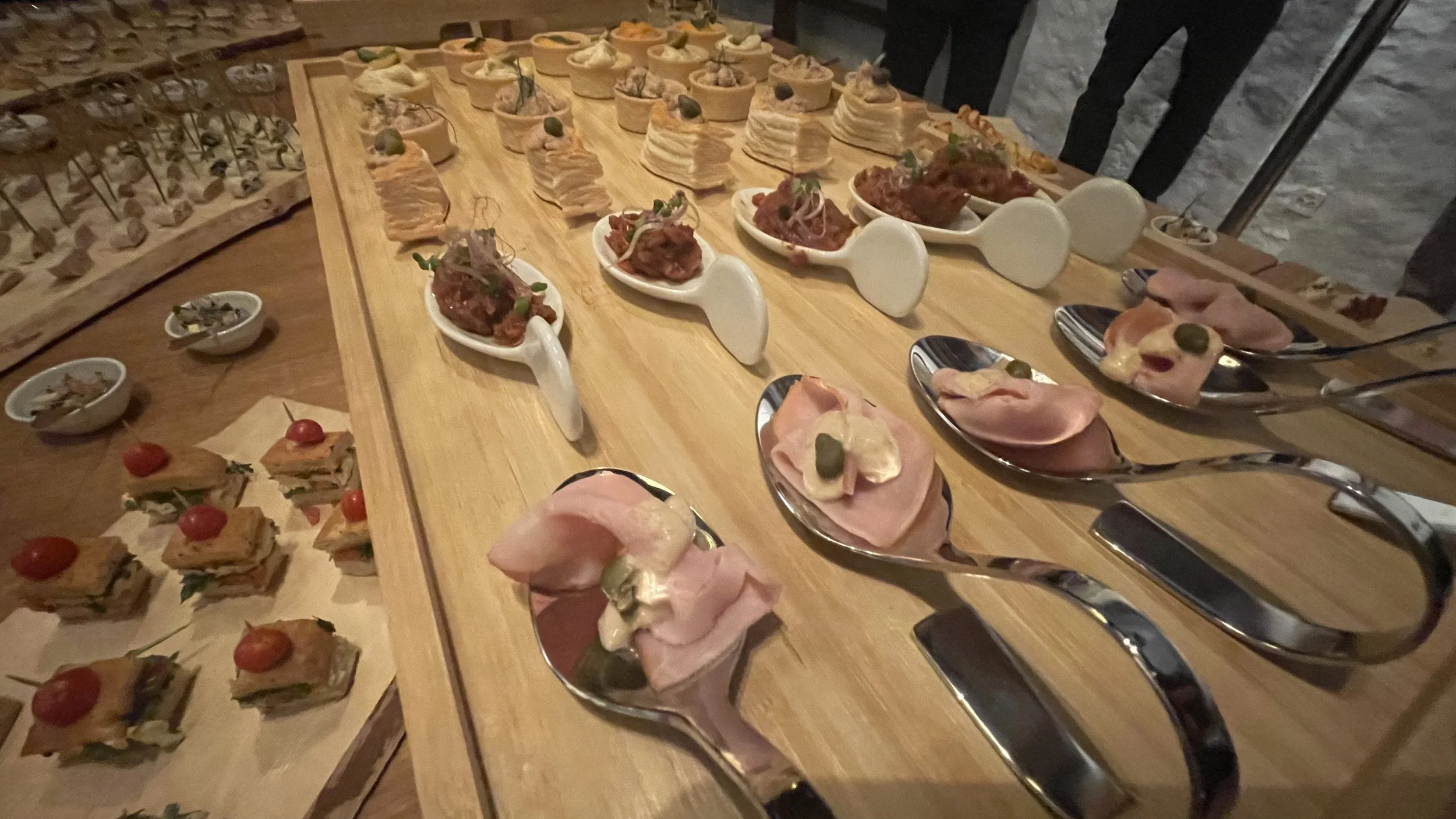 Verschiedene kleine Häppchen und Canapés auf einem Holztisch, inklusive Aufschnitt, kleine Sandwiches, Terrinen und Pasteten, serviert auf weißen Löffeln und silbernen Löffeln.