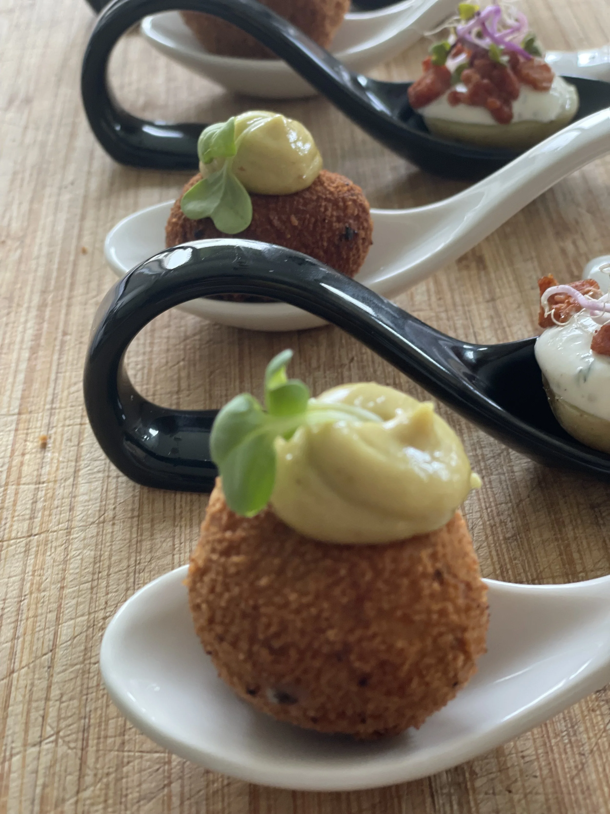 Verschiedene Canapés mit Cremes, Kräutern und knusprigen Beilagen auf schwarzen und weißen Löffeln auf einem holzigen Tisch.