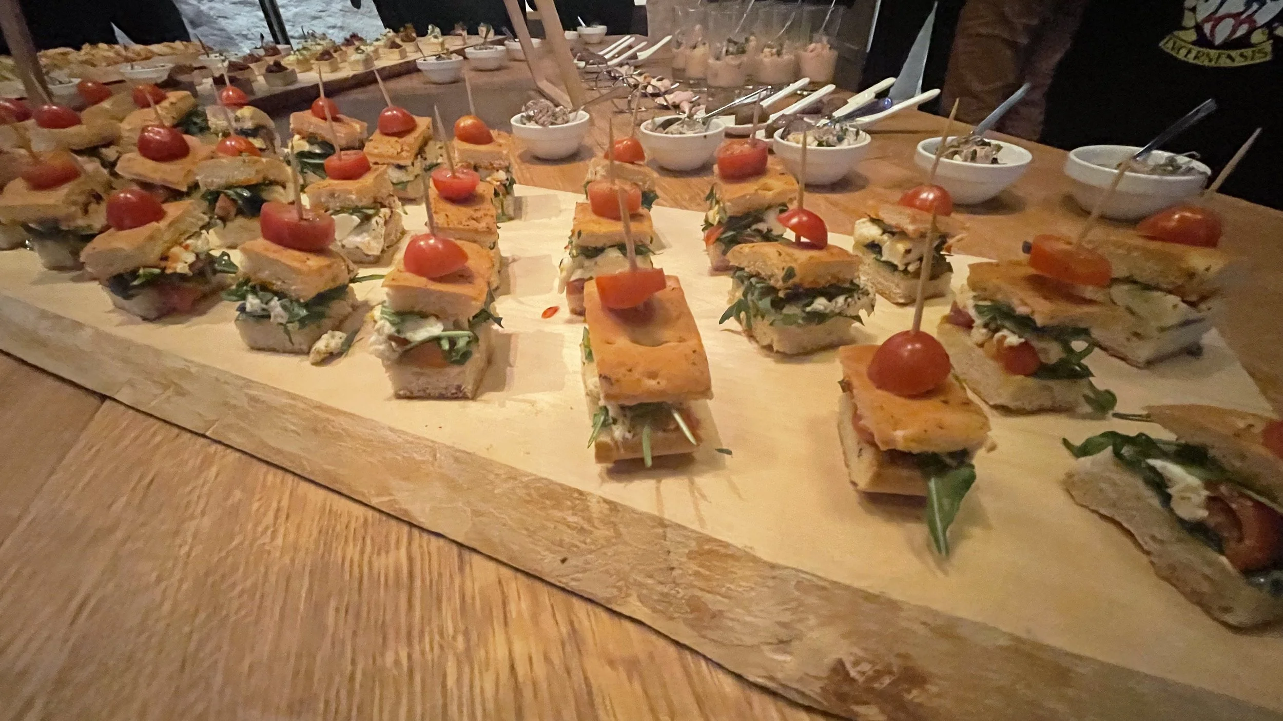Verschiedene kleine Sandwiches mit Tomaten, Salat und Käse auf Holzplatte, umgeben von Suppenschalen mit Dip