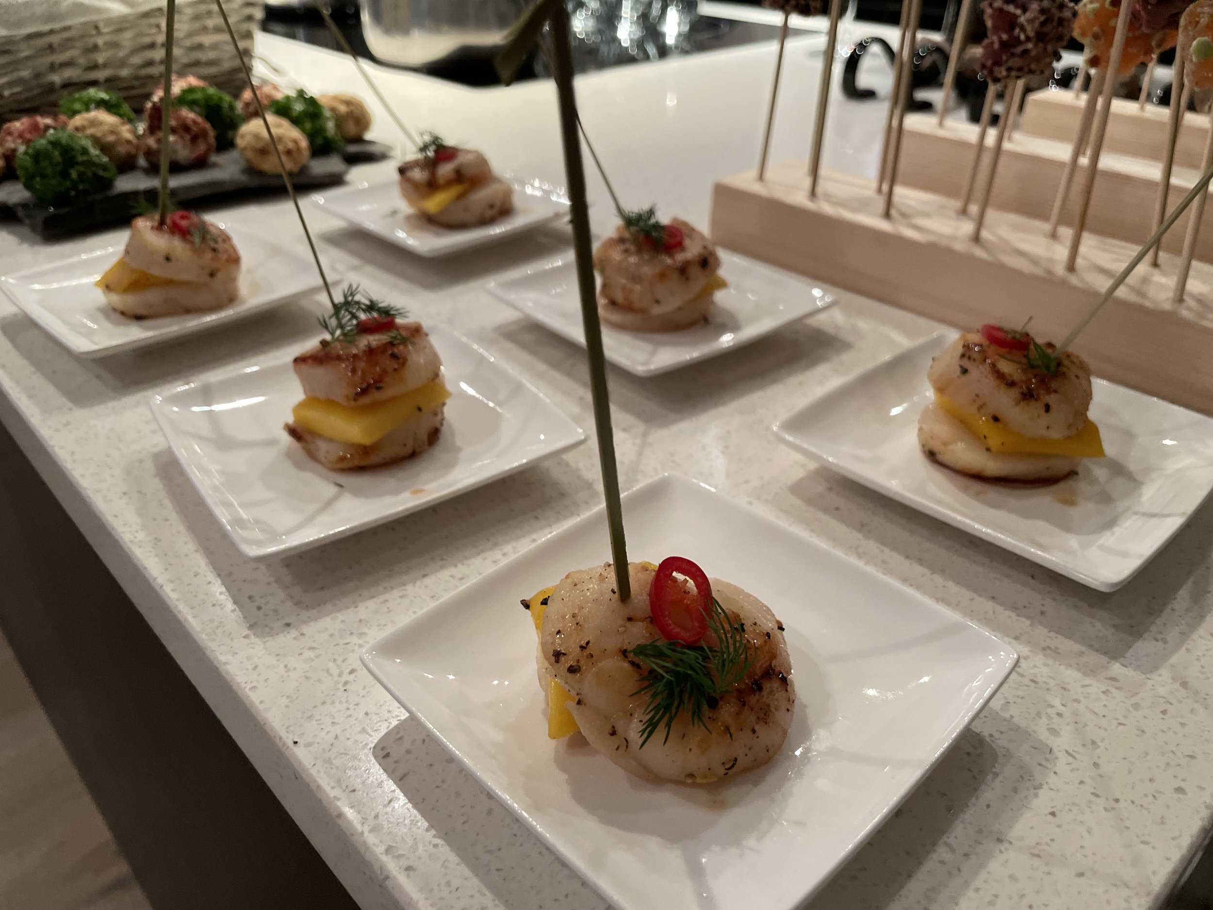 Verschiedene kleine Häppchen auf weissen Tellern, bestehend aus gegrilltem Fleisch, Käse, und Gemüse, dekoriert mit Kräutern und kleinen Paprikaringen, auf einem weissen Speisetisch bei einer Veranstaltung.