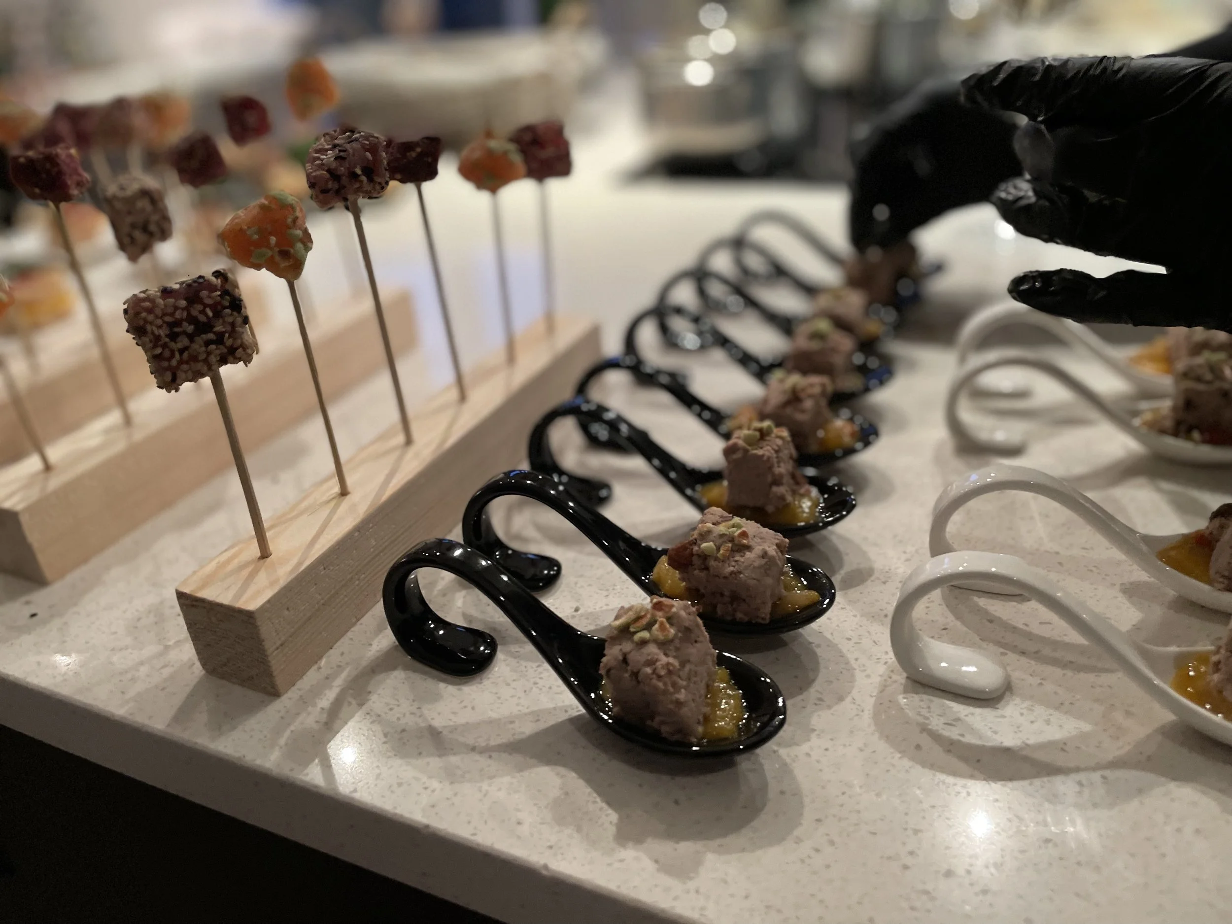 Verschiedene Häppchen und Snacks, präsentiert auf einem Buffet-Tisch, inklusive kleinen Löffeln mit Muscheln, schwarzen und weißen Löffeln, und Spießchen mit verschiedenen kleinen Speisen.