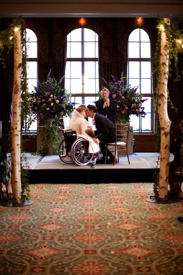The first kiss as a married couple - the bride in a wheelchair seated next to her husband - the celebrant leads the applause behind them