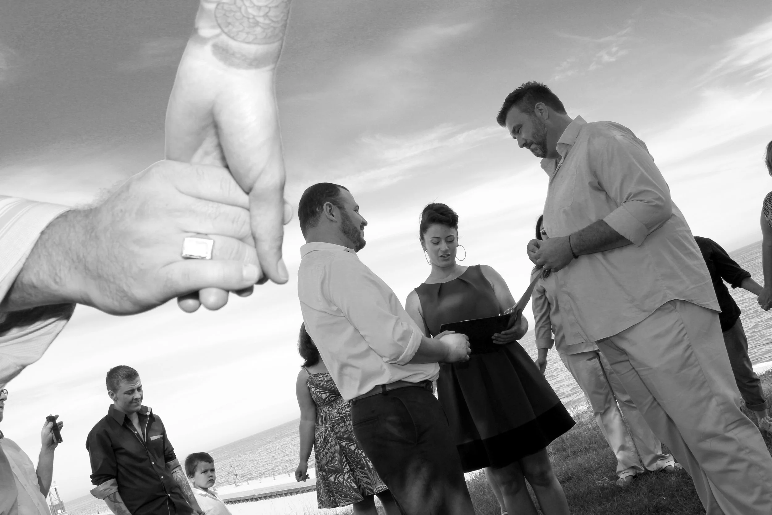 Matt and his partner Joel, both white men, are getting married. Their celebrant stands between them, she is holding a portfolio and reading from it. They are surrounded by their family and friends in a circle holding hands.