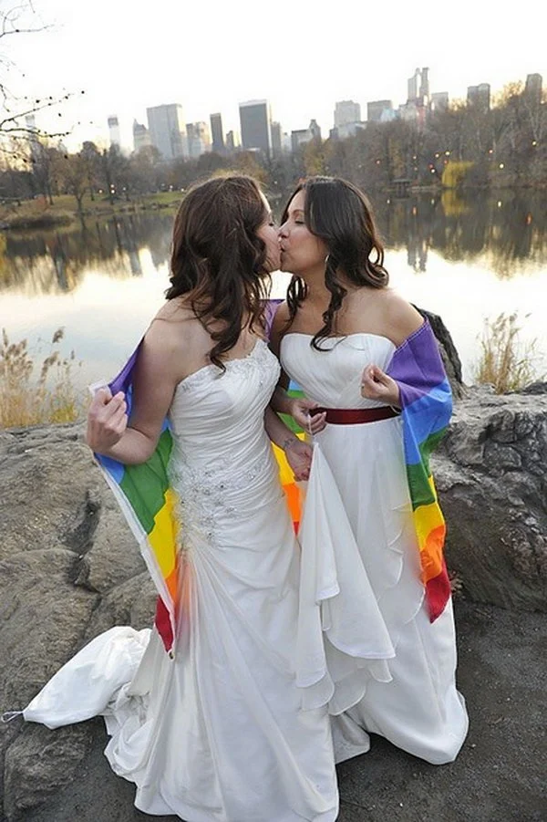 Two brides in white dresses share a kiss wrapped in a rainbow flag