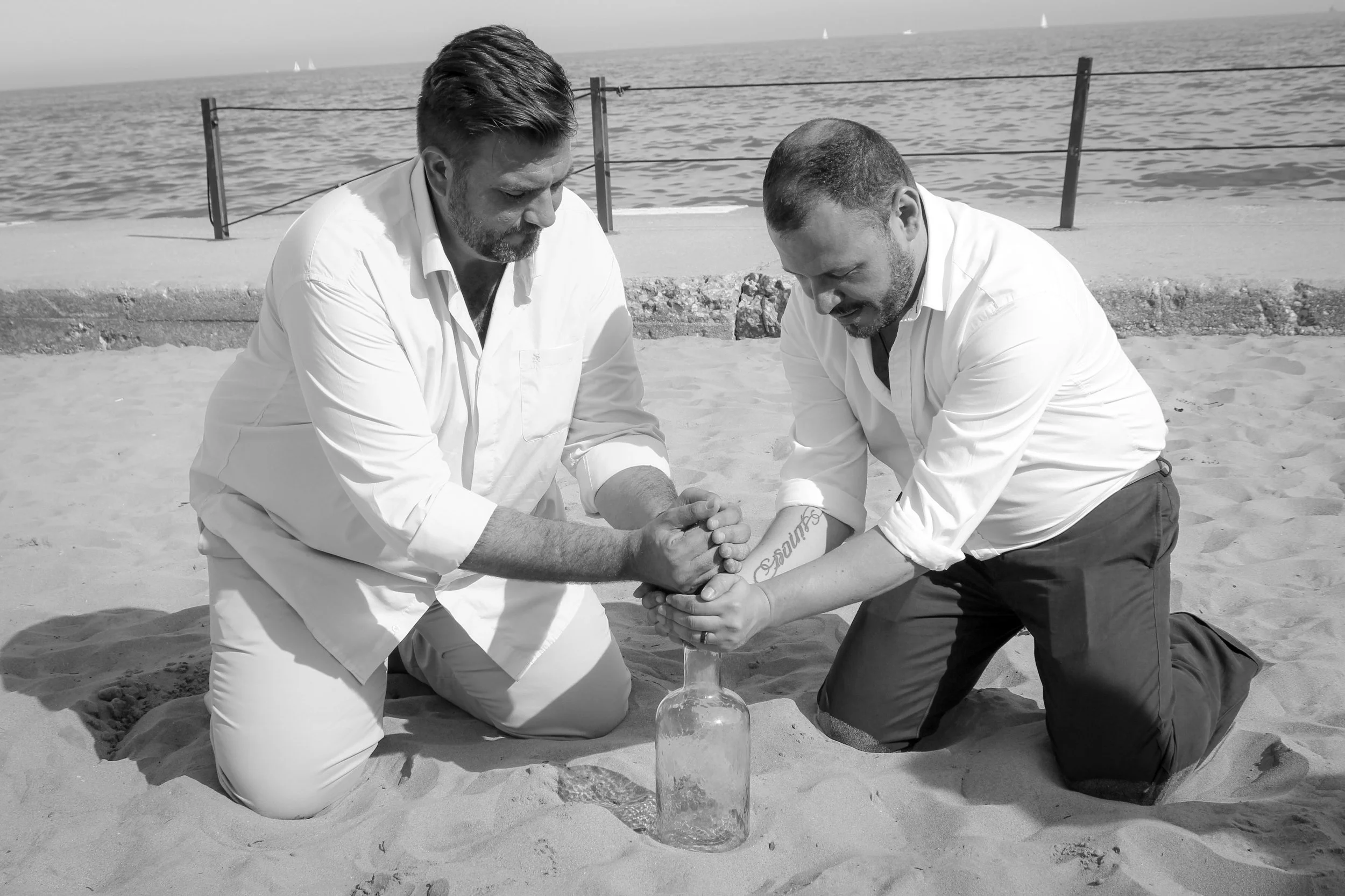 Two grooms kneeling on the beach, fill a bottle with sand