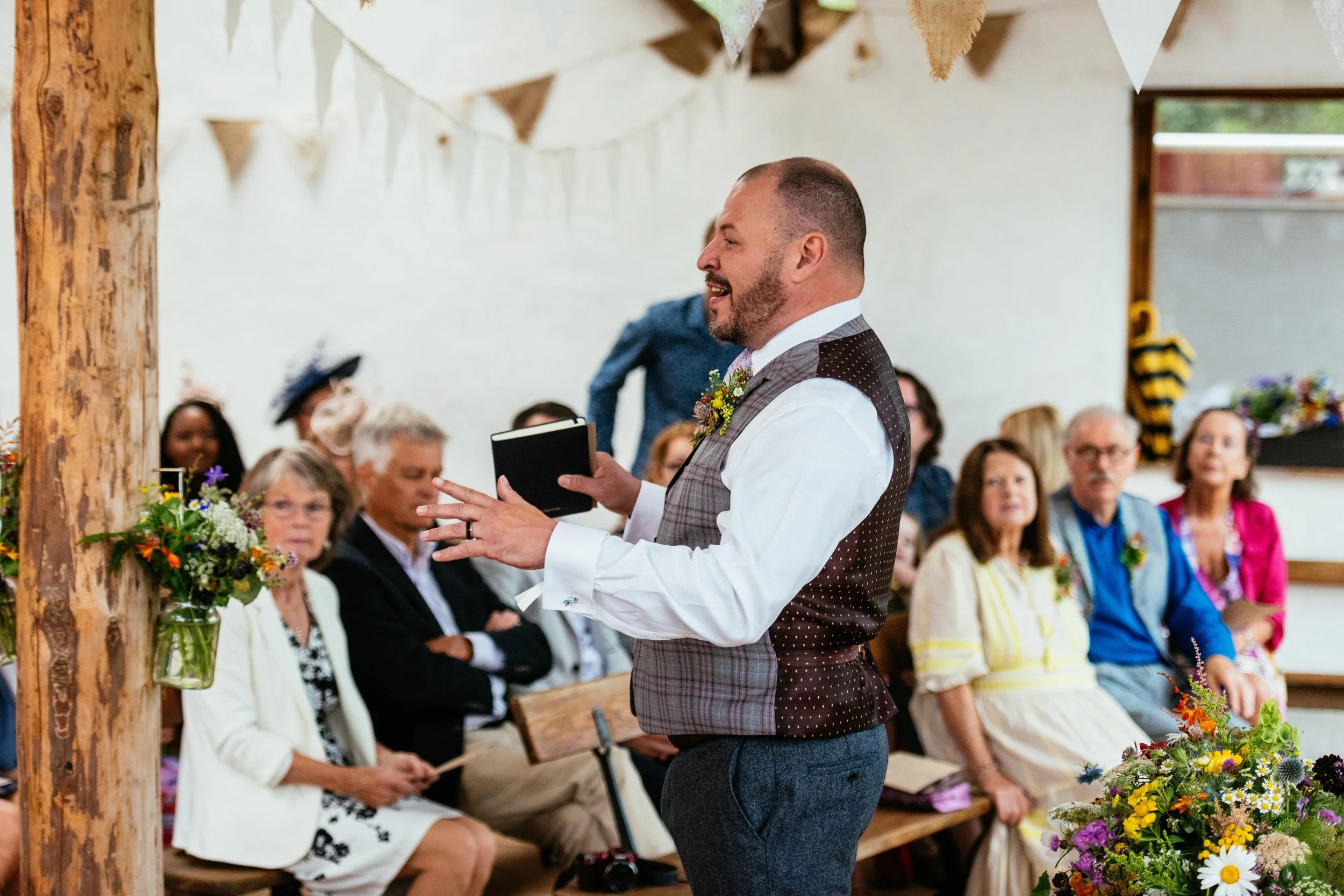 Matt is mid-flow talking about housekeeping before the couple arrives. He is a white middle aged man in a waistcoat, shirt and trousers and his hands are moving as he explains the order of service.