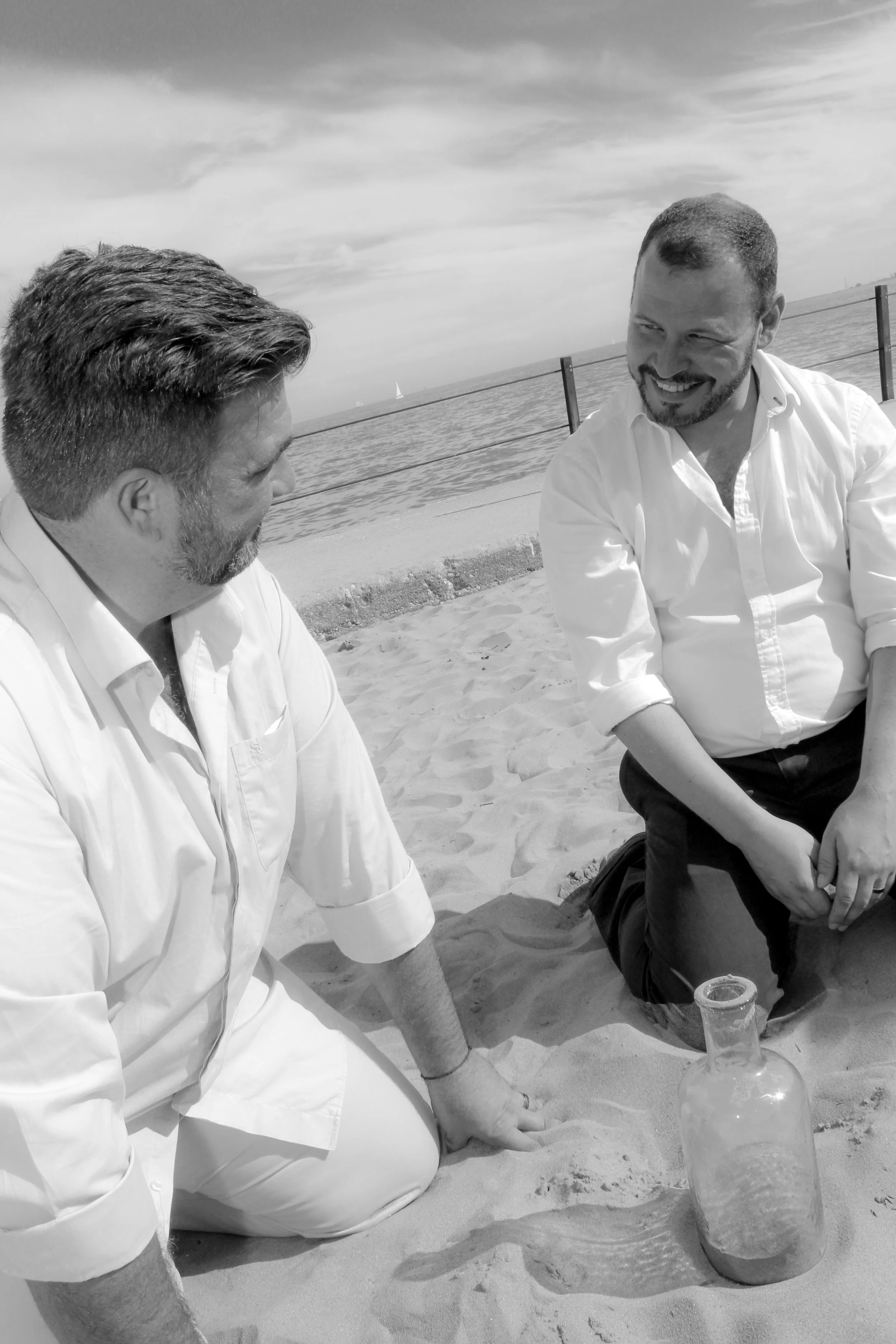 Matt and Joel, both white men in their late 30s are kneeling in the sane with a large glass bottle between them. They are smiling at one another. The bottle has some sand in it that they have just poured in.