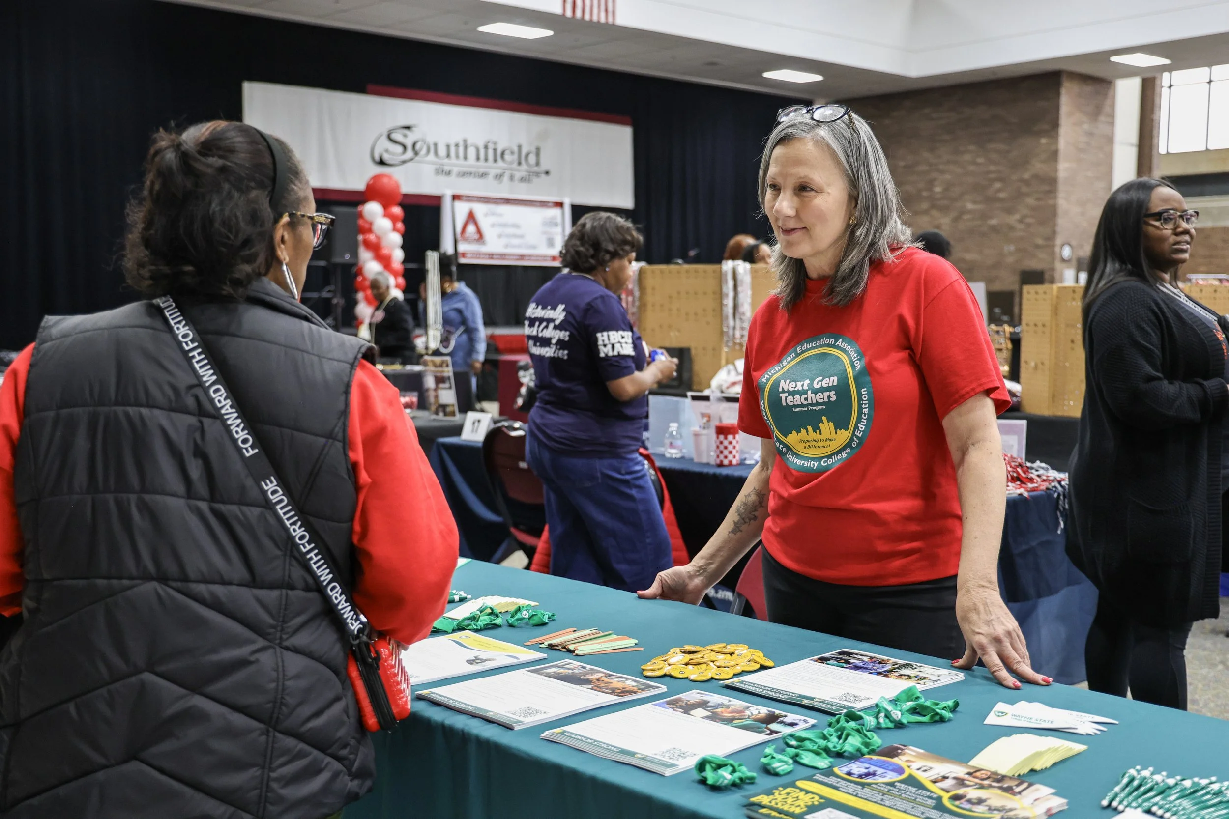 A woman with gray hair wearing a red T-shirt discussing with another woman at an informational booth, with several other booths and people in the background, in an indoor event space.
